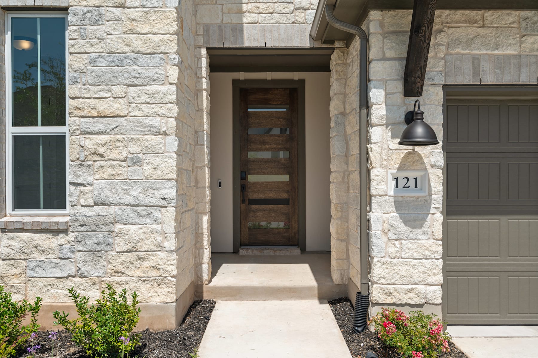 A stone-clad entryway with a wooden door, a paved walkway, and a lamp fixture, surrounded by lush greenery and flowers.