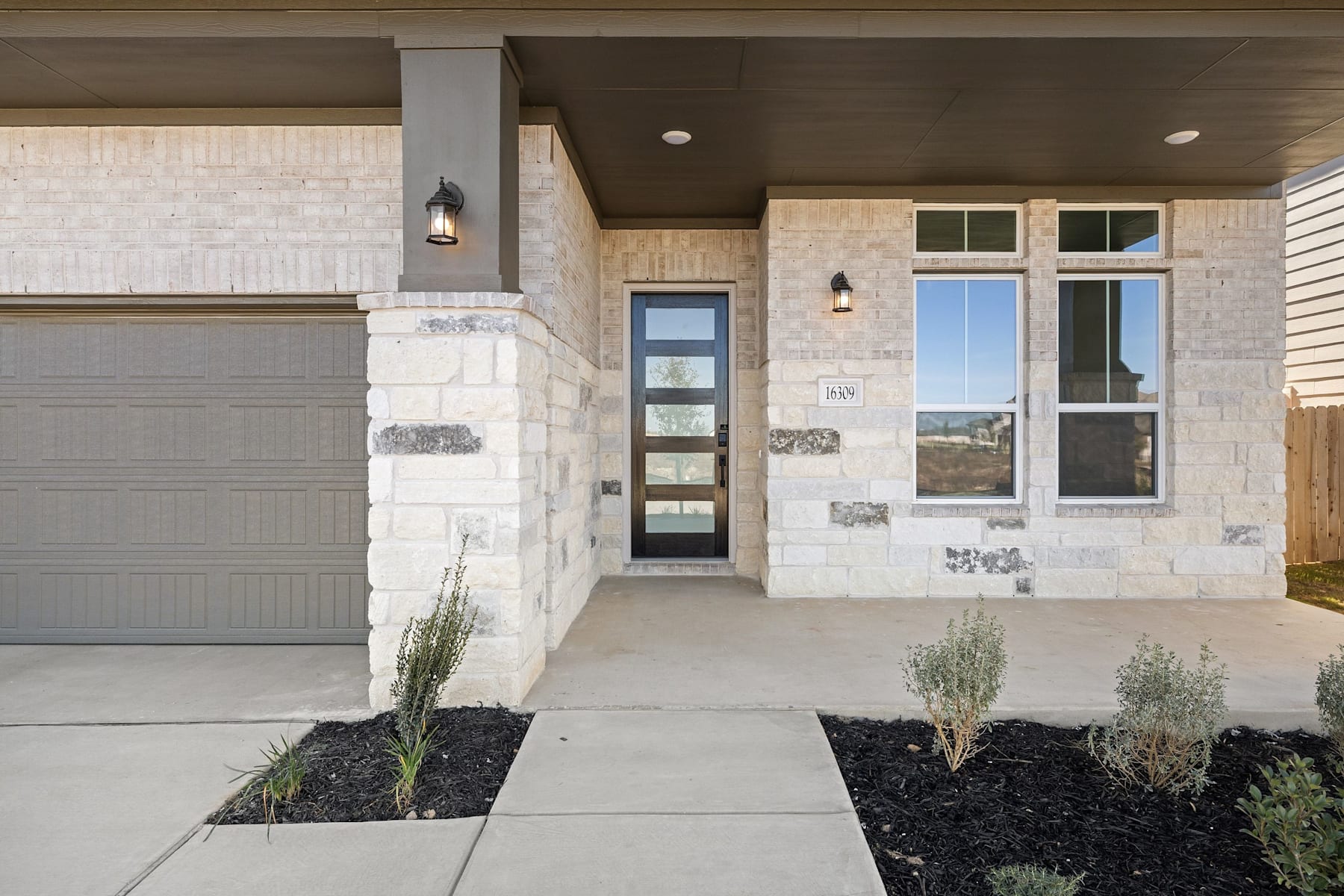 A modern, two-story house with a stone exterior, a garage door, and a landscaped front yard with plants and a concrete walkway leading to the entrance.