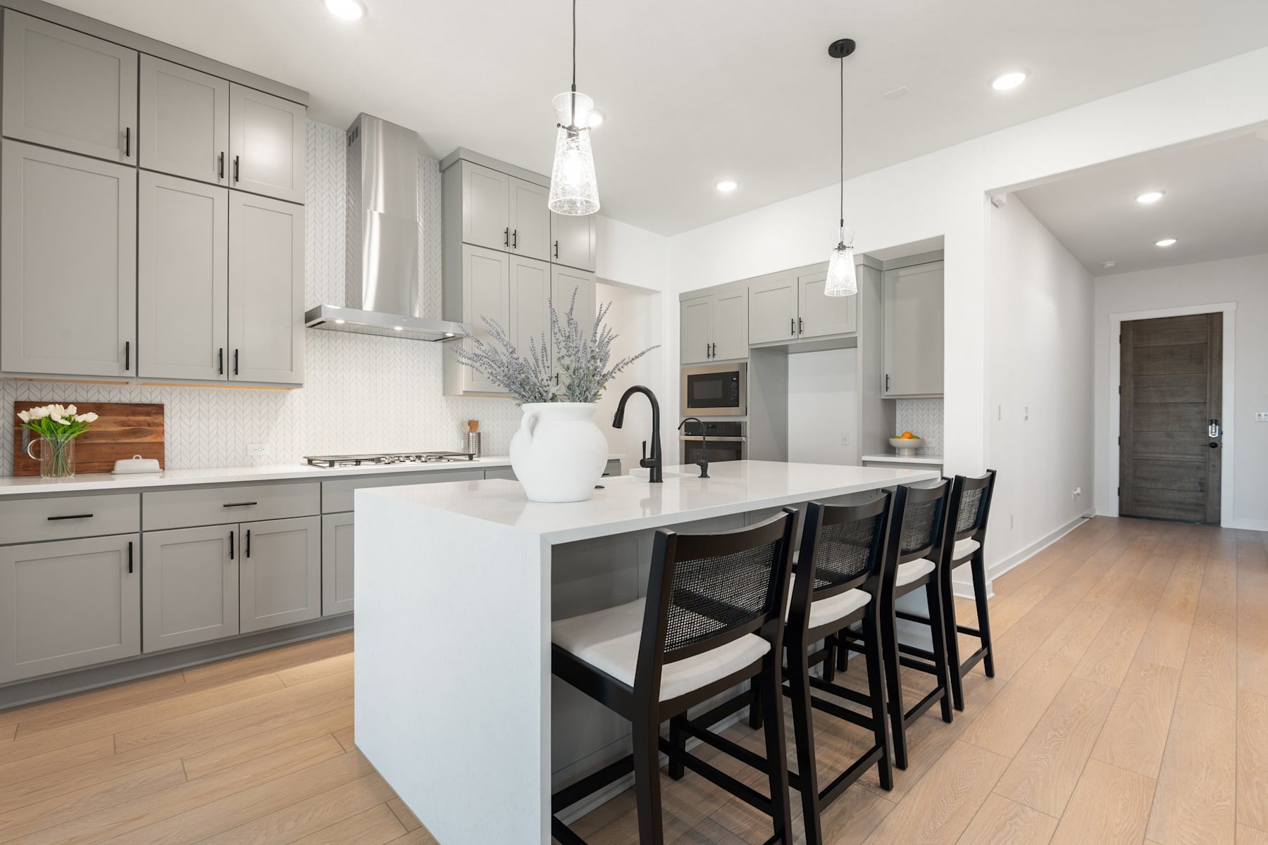 A modern, minimalist kitchen with gray cabinets, a white countertop, and black bar stools, complemented by pendant lights and a hardwood floor.