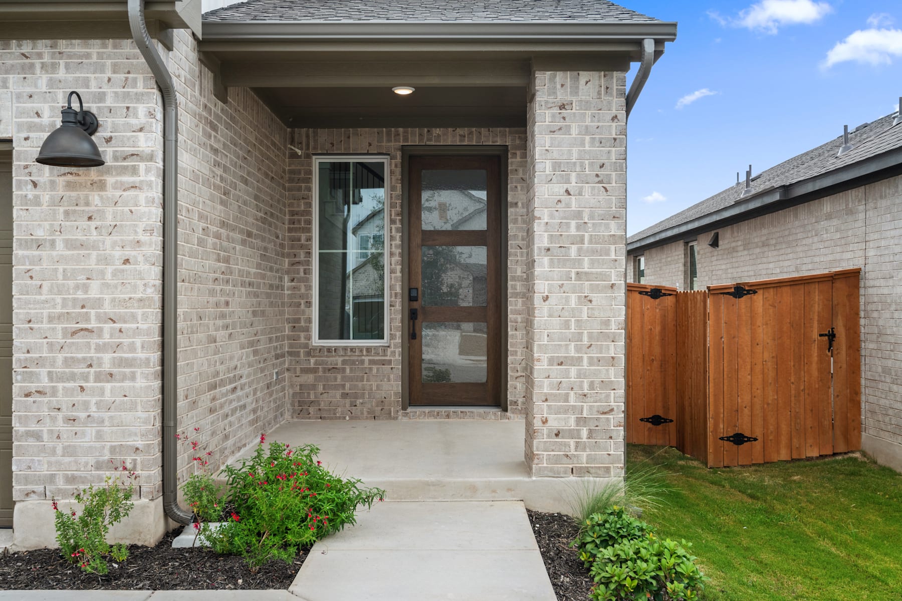 A brick house with a wooden front door, a porch, and a grassy yard with plants in the foreground.