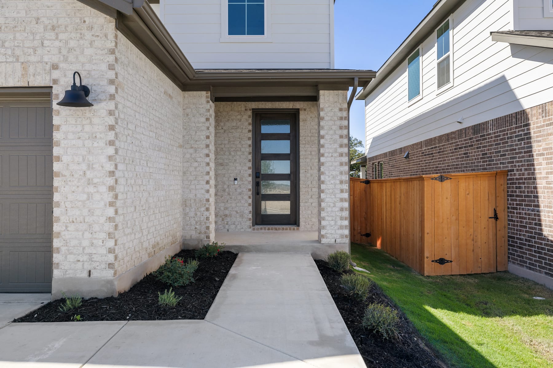 A modern, well-designed residential home with a paved walkway leading to the front entrance, surrounded by landscaped greenery and a wooden fence in the background.