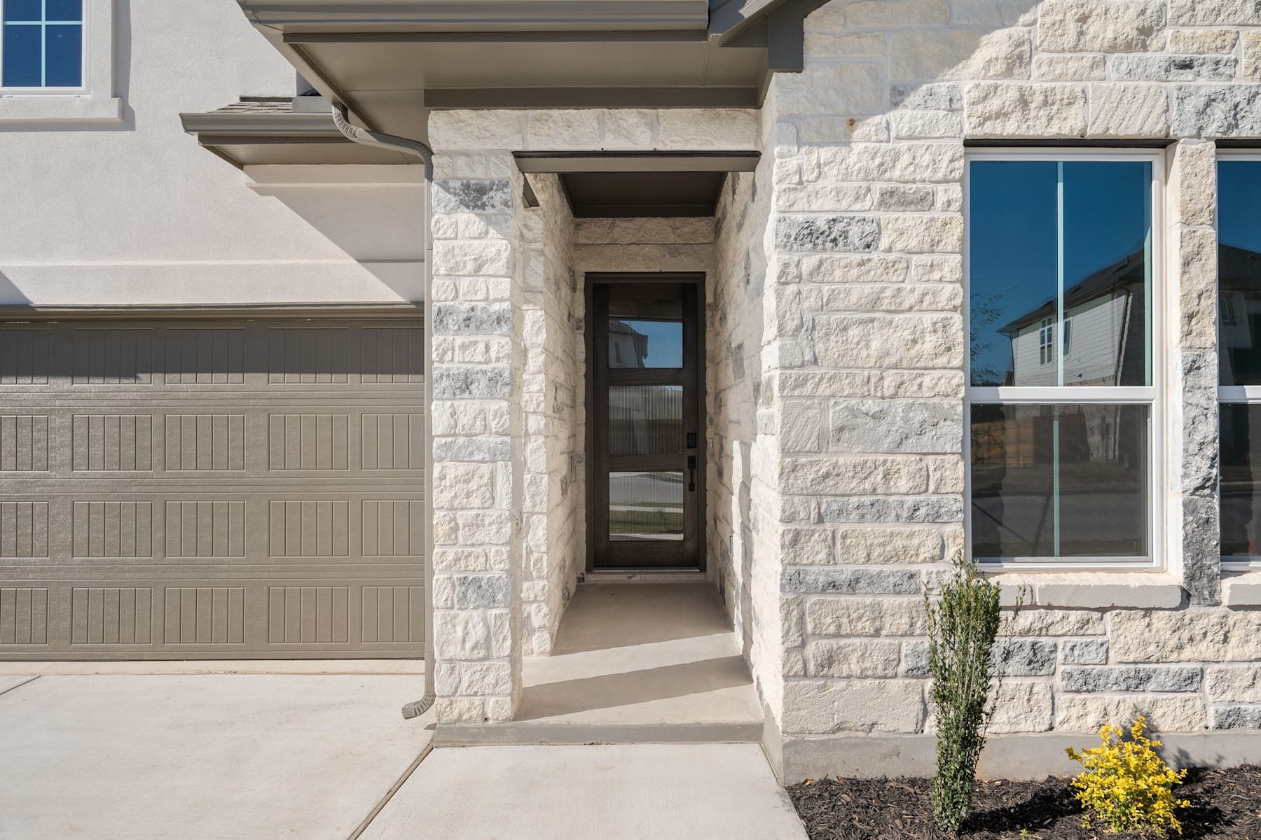 A modern, two-story residential building with a stone exterior, a covered entryway, and a garage door in the foreground. The building is surrounded by landscaping, including a small plant or shrub in the foreground.