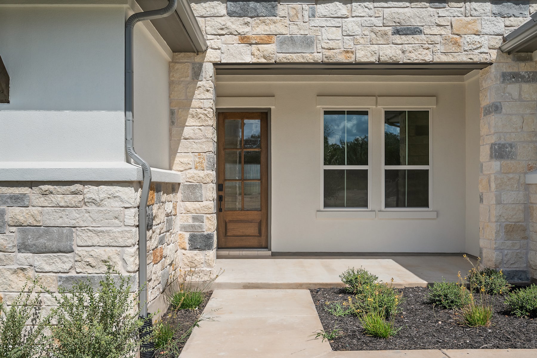 A stone-clad entryway with a wooden door and windows, surrounded by landscaped greenery, leading to a modern residential structure.