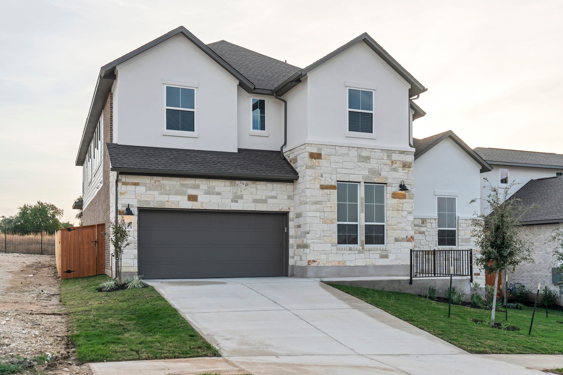 A two-story house with a gray roof, white siding, and stone accents, surrounded by a grassy lawn and a concrete driveway leading to a garage door.
