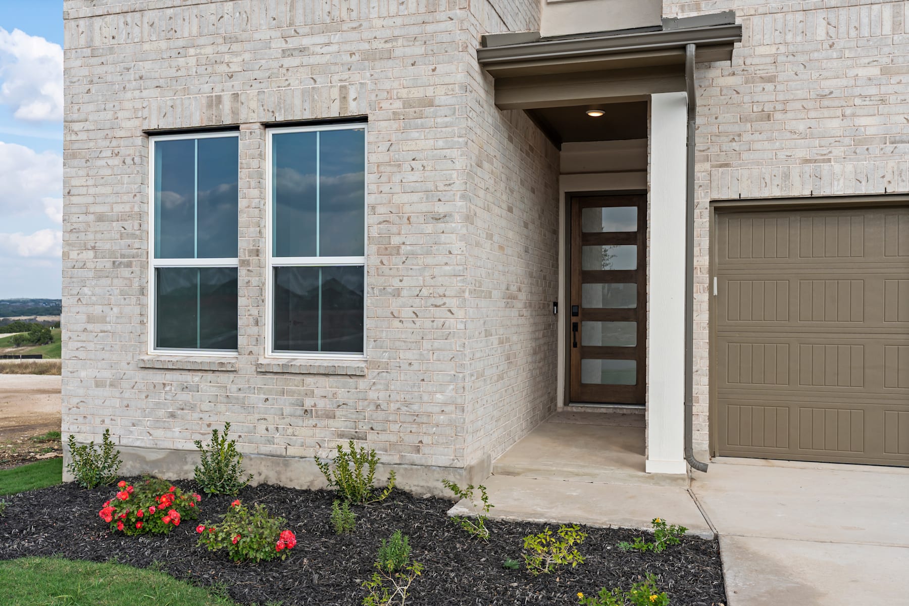 A modern, two-story residential building with large windows, a wooden front door, and a well-landscaped yard featuring colorful flowers and a grassy area in the foreground.