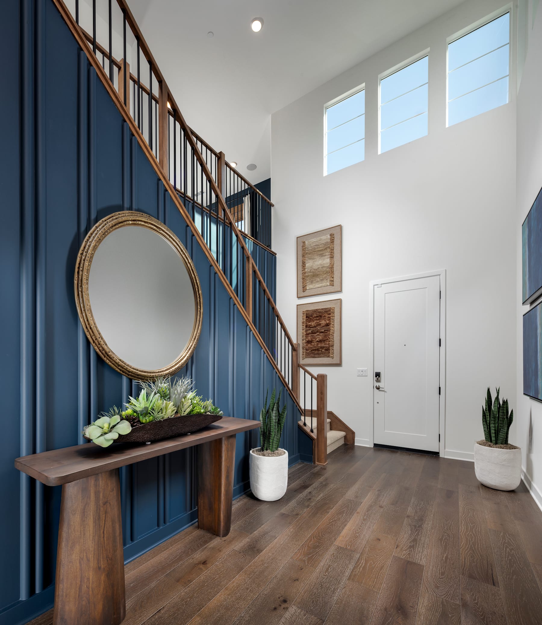 A spacious and well-lit entryway with a wooden bench, a round mirror, and potted plants, set against a backdrop of a staircase with a navy blue accent wall.