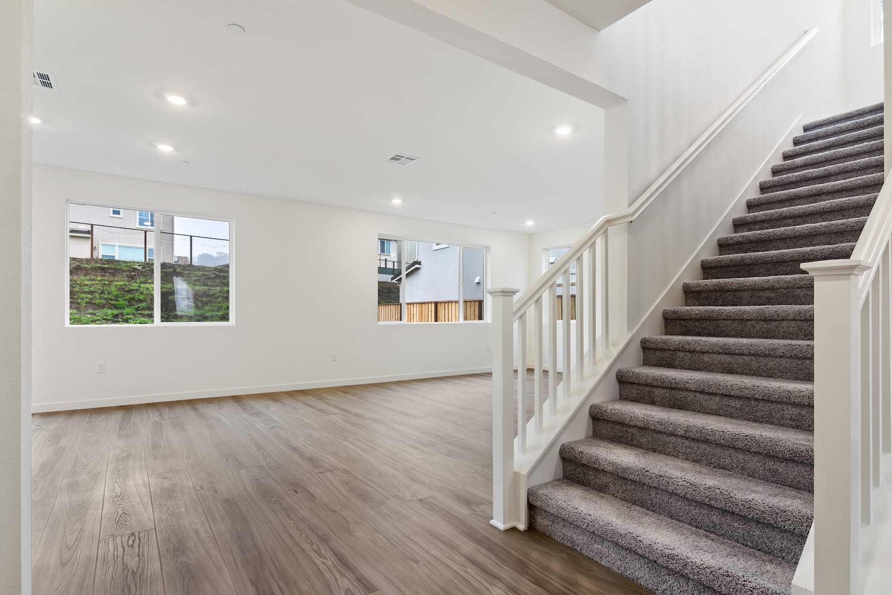 A spacious and bright interior with a wooden floor, a staircase with carpeted steps, and a window overlooking a green outdoor area.
