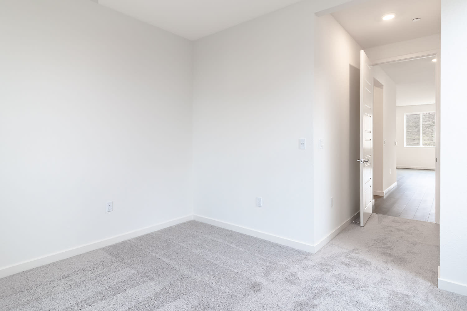 A bright, minimalist room with white walls, a gray tiled floor, and a doorway leading to a hallway.