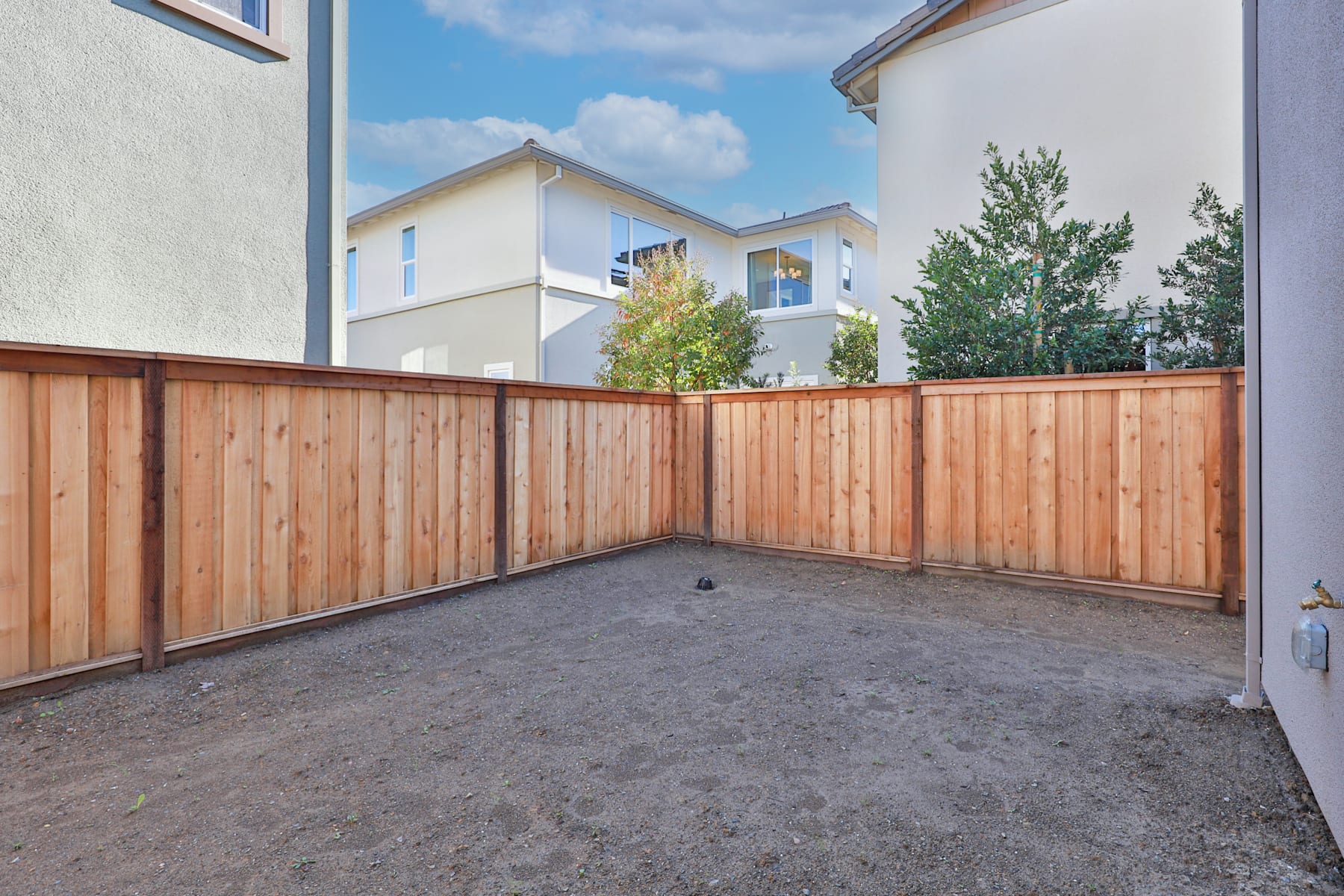 A wooden fence surrounds a paved area in the foreground, with residential buildings and trees visible in the background under a cloudy blue sky.