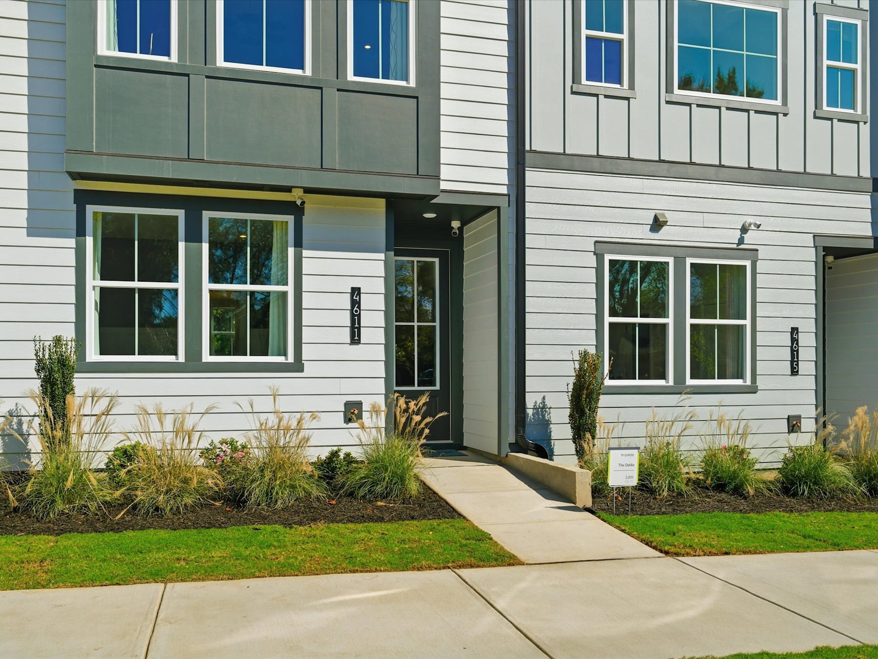 A modern, multi-story residential building with gray siding, large windows, and a paved walkway leading to the entrance, surrounded by landscaped greenery.