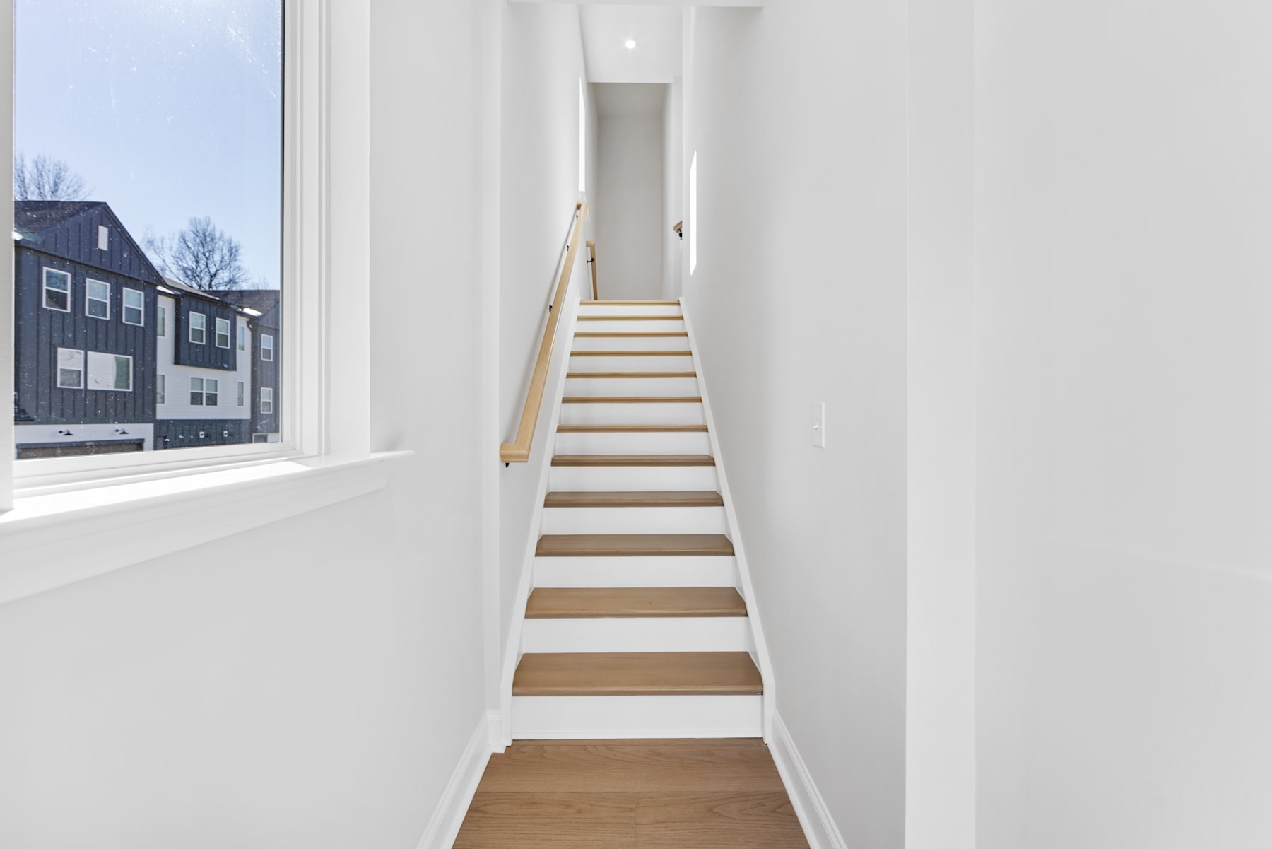 A bright, minimalist staircase leads up from a wooden floor, with a large window in the background revealing a view of buildings outside.