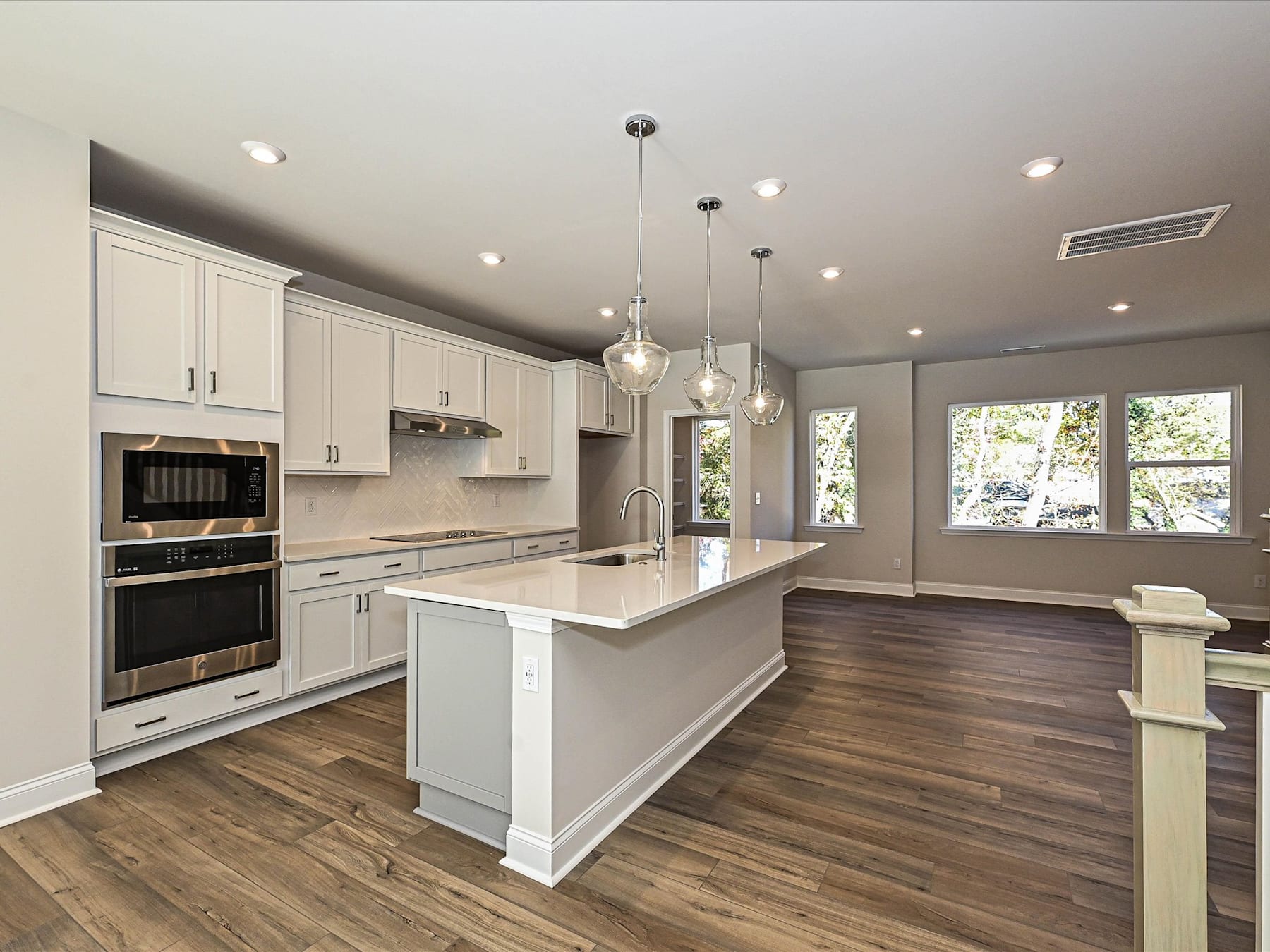 A modern, open-concept kitchen with white cabinets, stainless steel appliances, and a large central island with a marble-like countertop, set against a backdrop of large windows overlooking a wooded outdoor area.