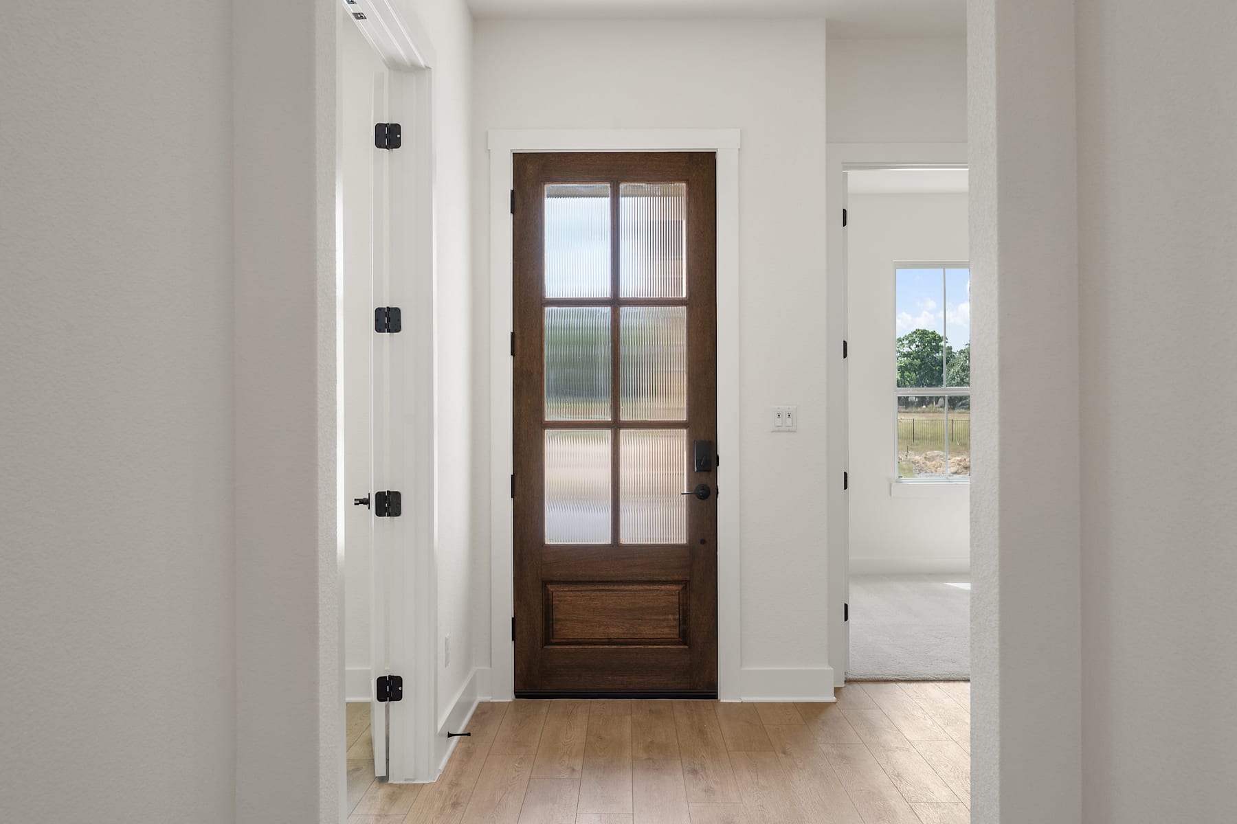 A hallway with a wooden door featuring a glass panel, leading to a bright and open space beyond.