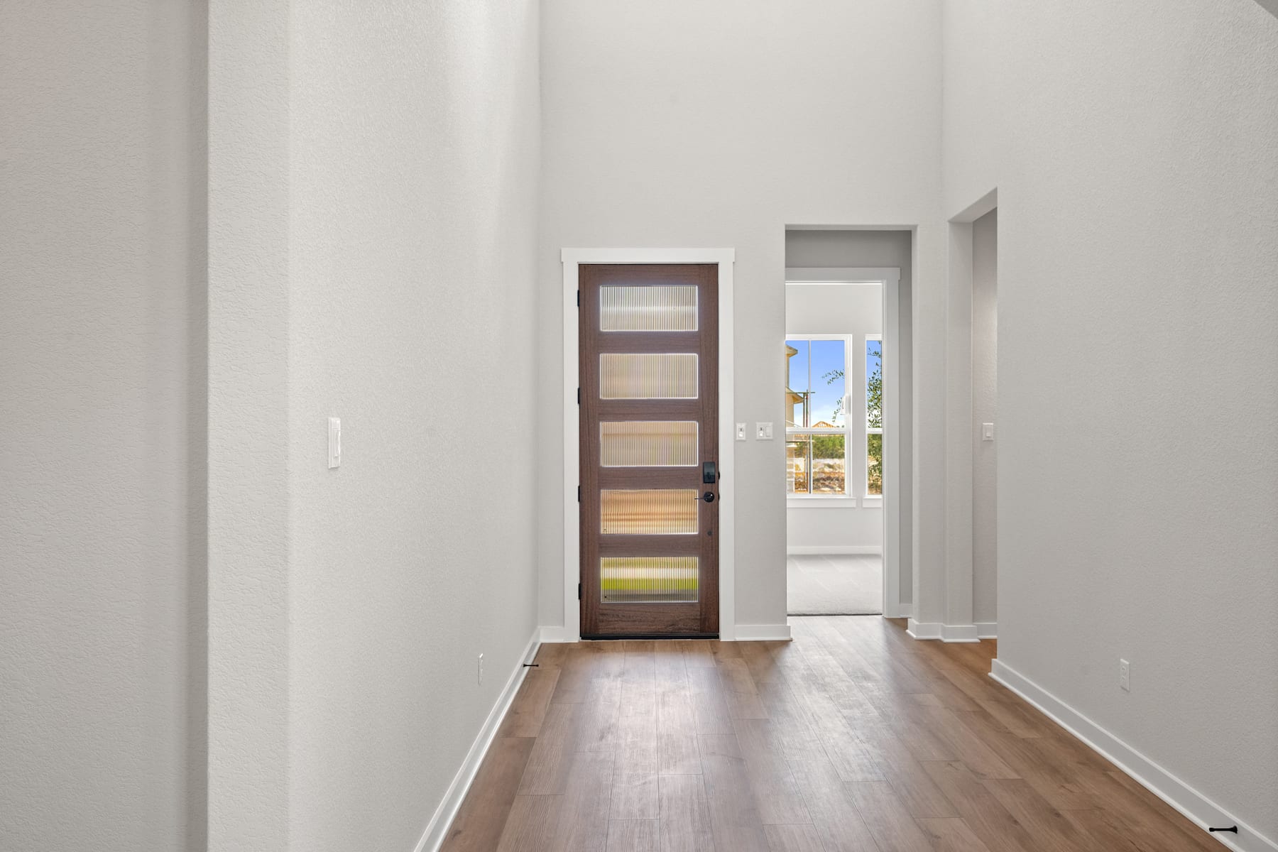 A hallway with a wooden door leading to an outdoor view, featuring a hardwood floor and white walls.
