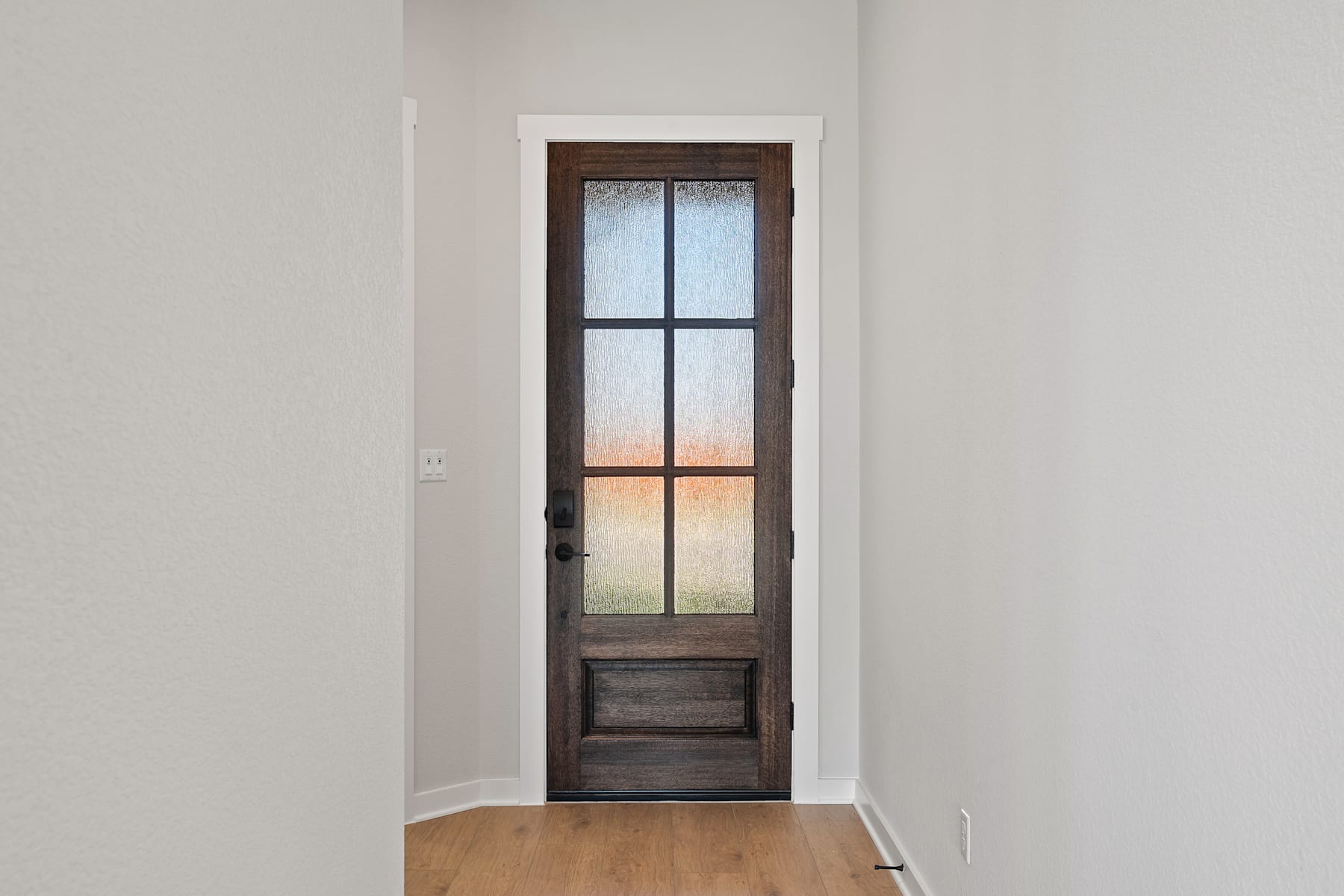 A wooden door with a glass panel stands in a white-walled room, leading to what appears to be another space beyond.