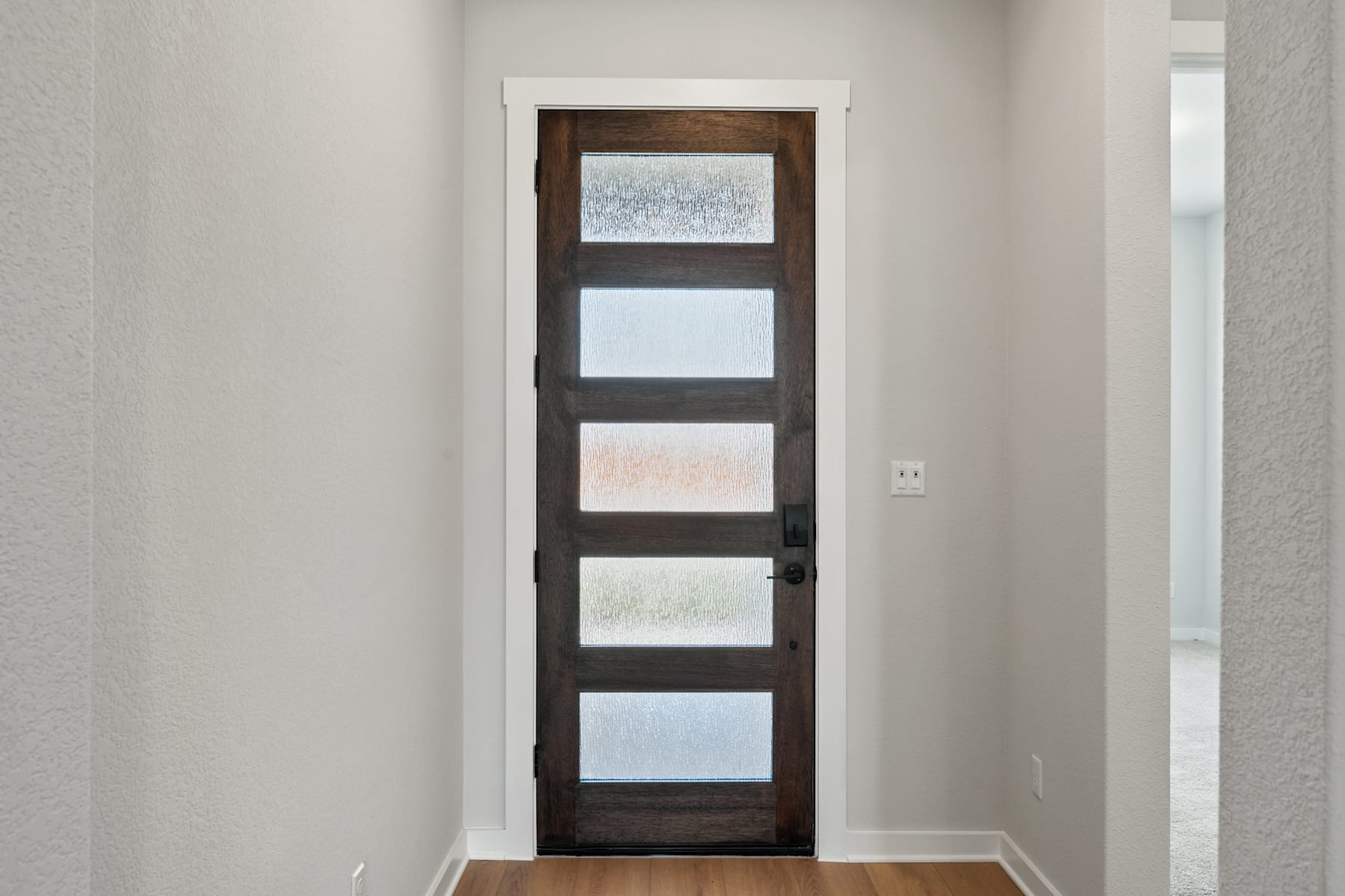 A wooden door with frosted glass panels stands in a bright, minimalist room with white walls.
