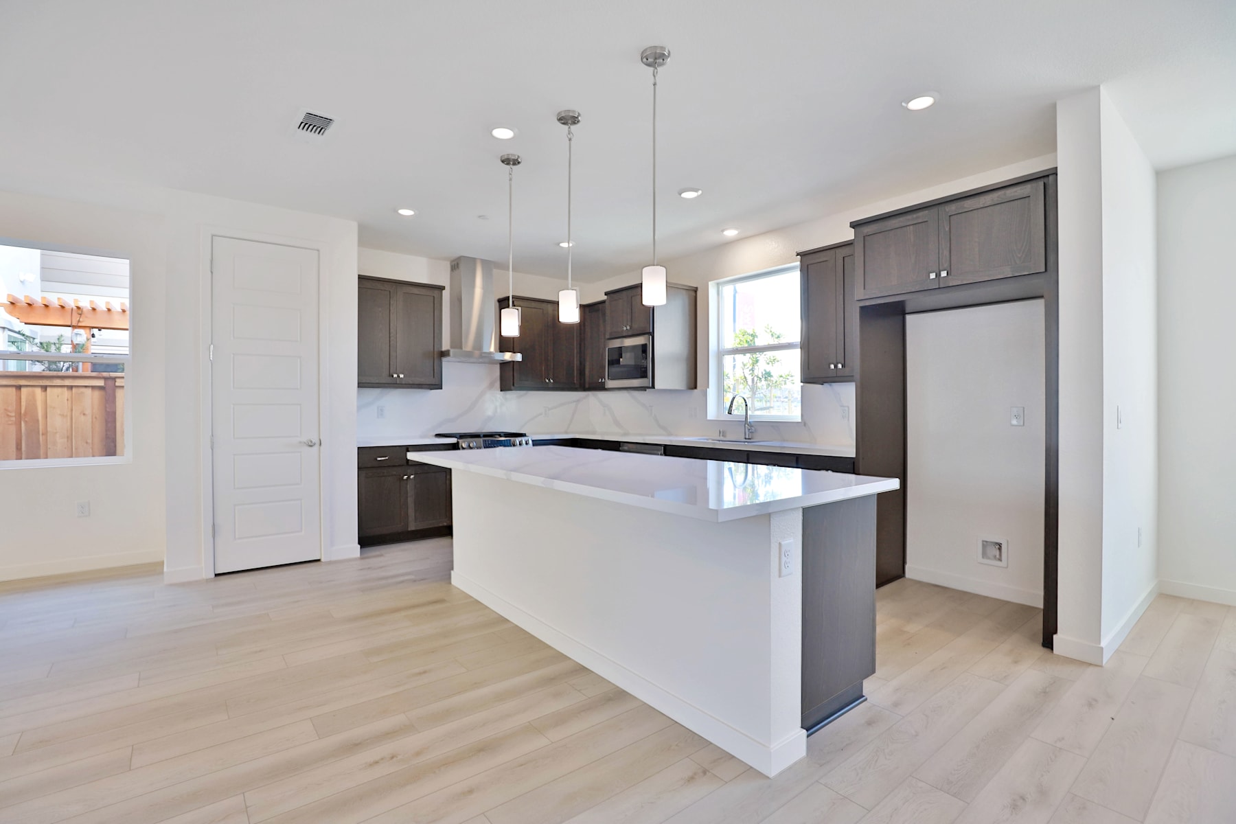 A modern, open-concept kitchen with dark cabinets, a white countertop, and pendant lighting fixtures, set against a light-colored hardwood floor.