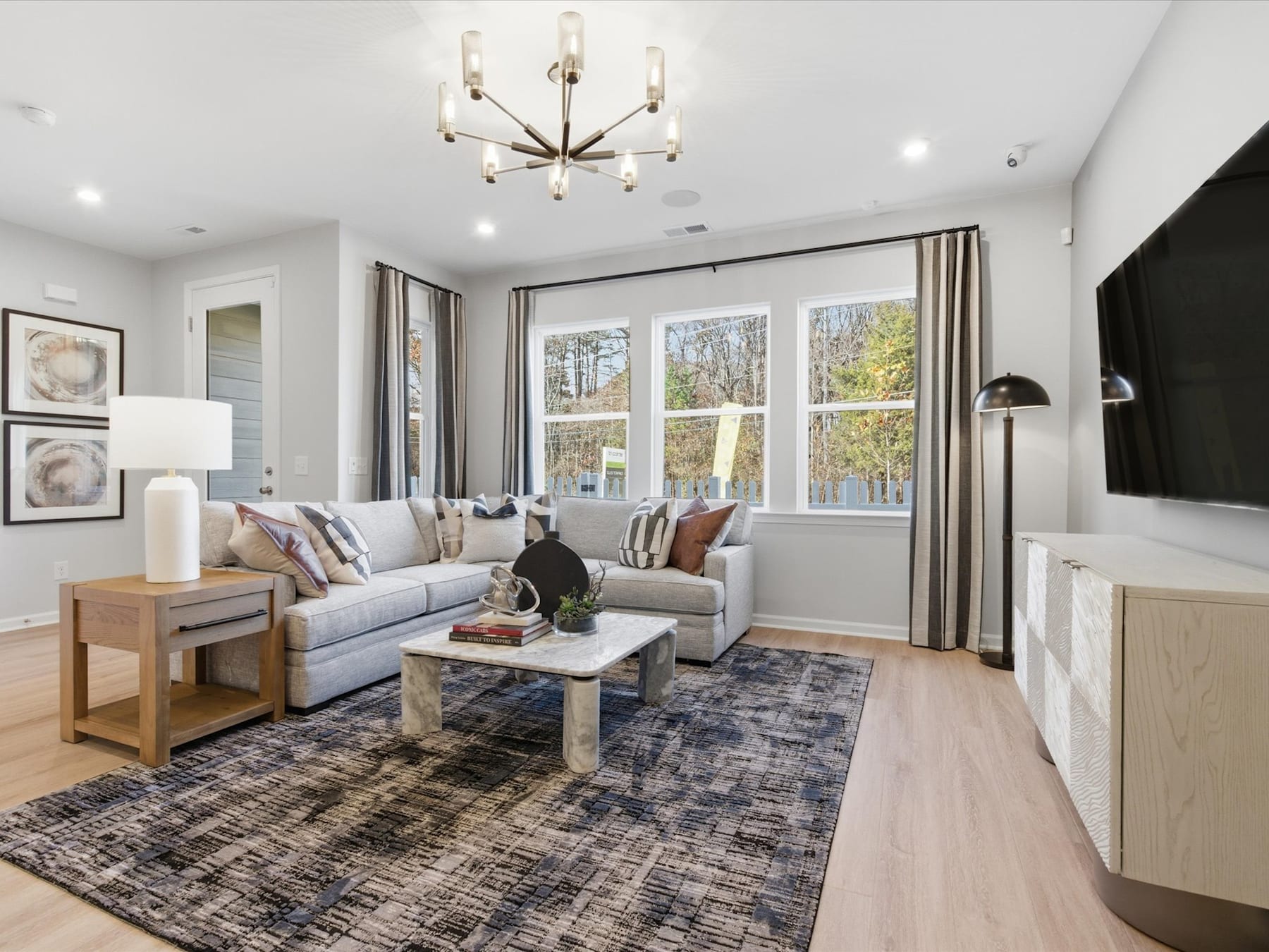 A cozy and well-furnished living room with a gray sectional sofa, a wooden coffee table, and a modern chandelier hanging from the ceiling, surrounded by large windows that provide natural light and a view of the outdoor scenery.