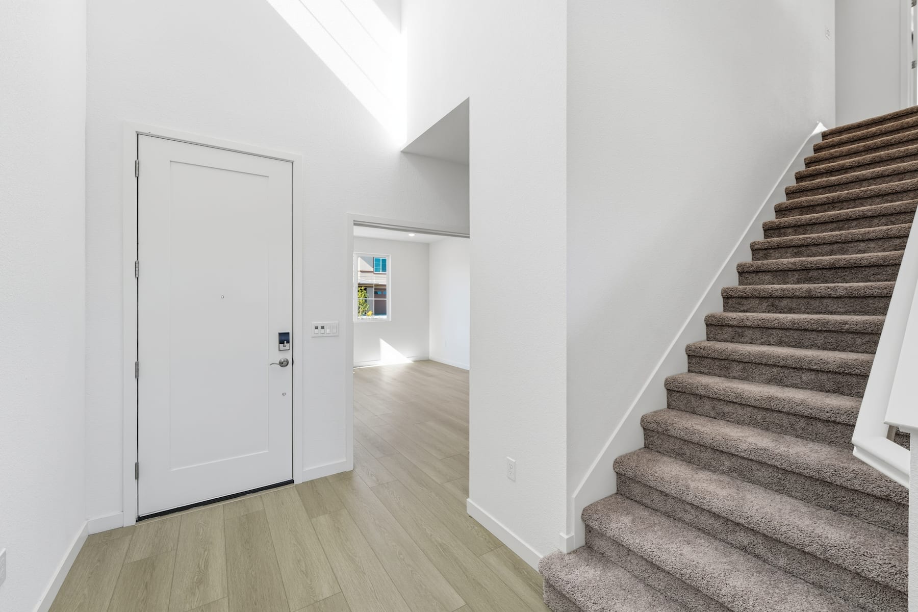 A bright and spacious entryway with a white door, wooden stairs, and a hardwood floor leading to a well-lit hallway.