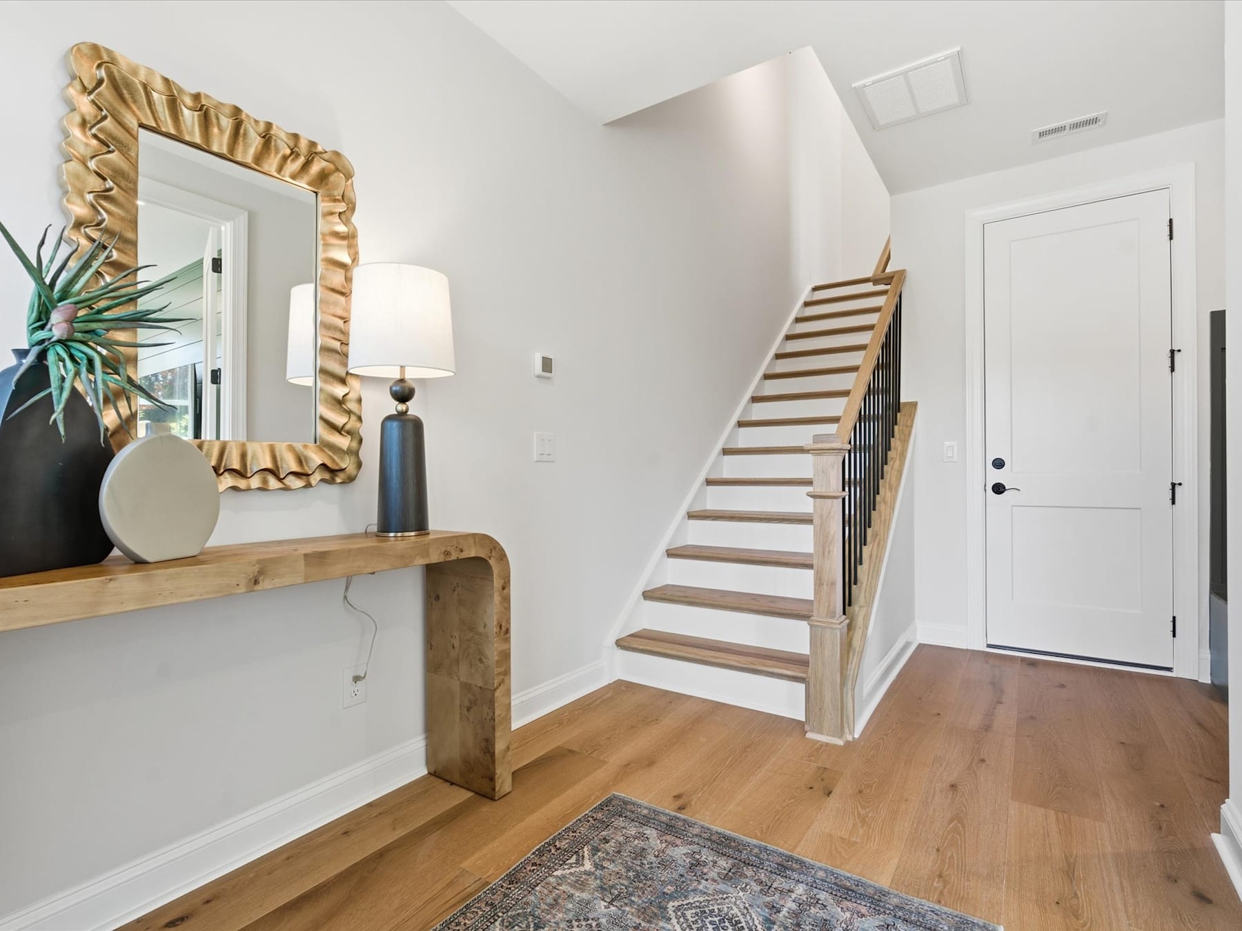 A spacious and well-lit entryway with a wooden staircase, a decorative mirror, and a wooden console table adorned with vases and plants.