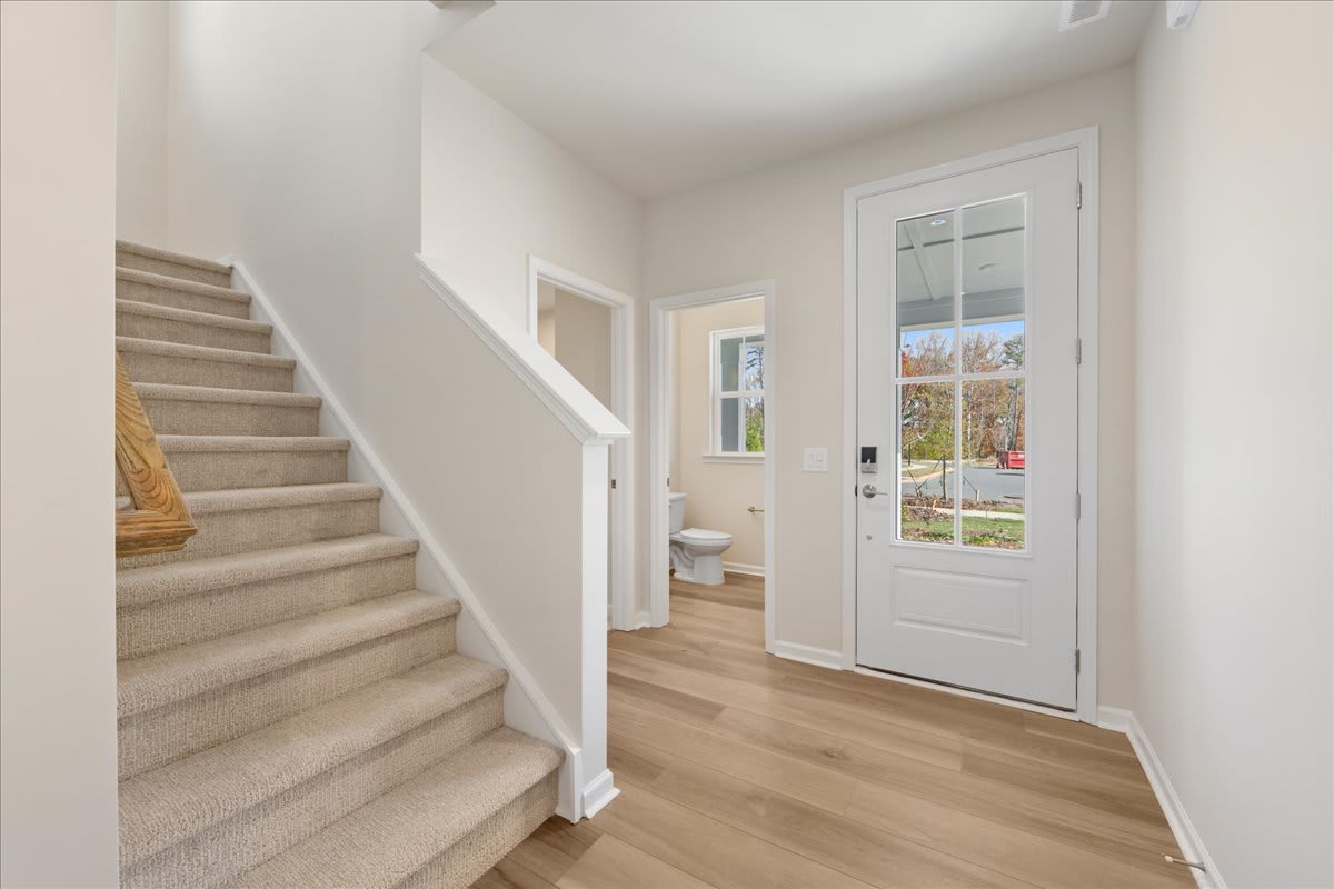 A bright and airy entryway with a carpeted staircase leading up, a white door with a window, and a hardwood floor in the background.