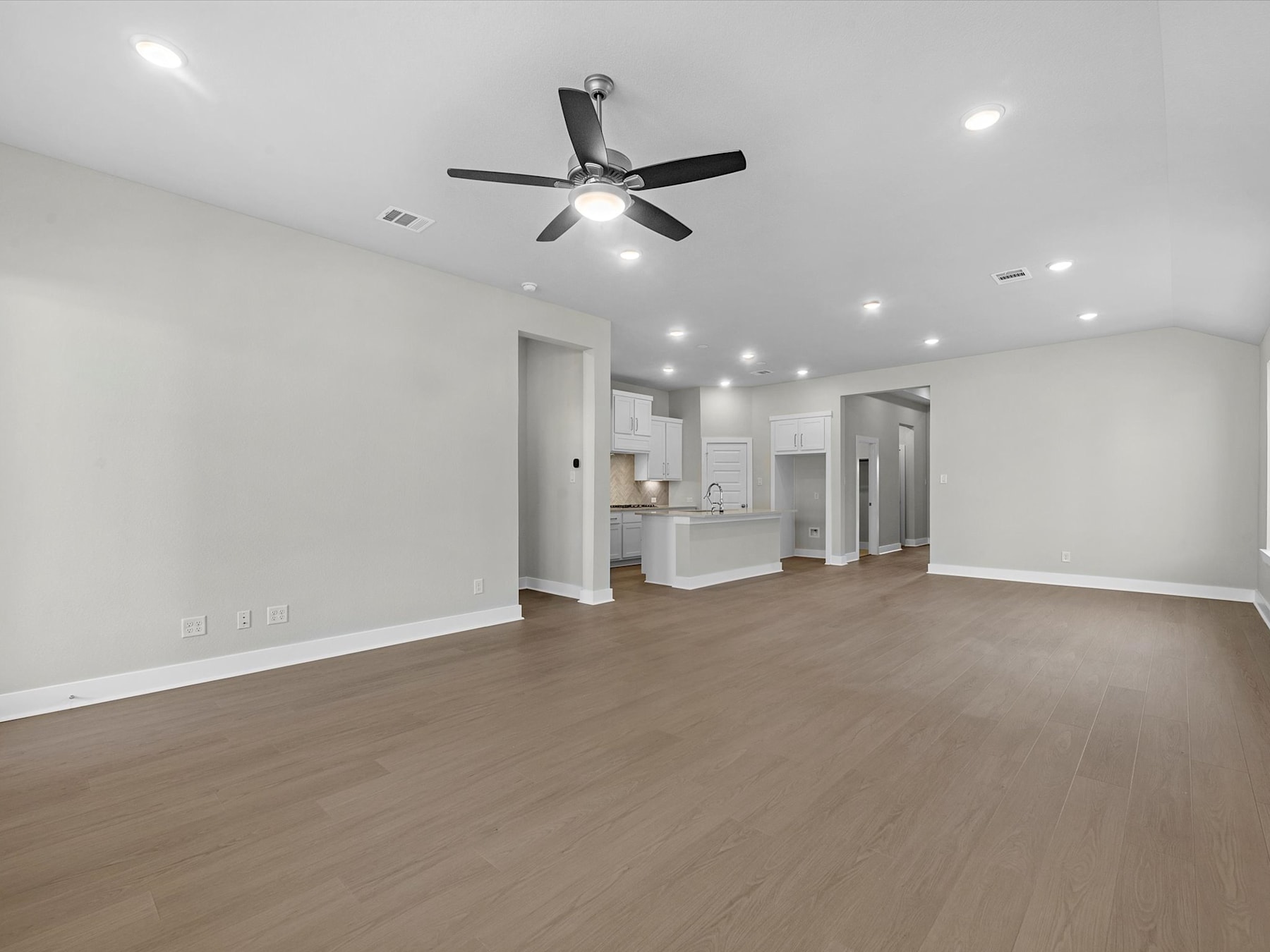 A spacious, open-concept room with a ceiling fan, hardwood floors, and a view of a kitchen area in the background.
