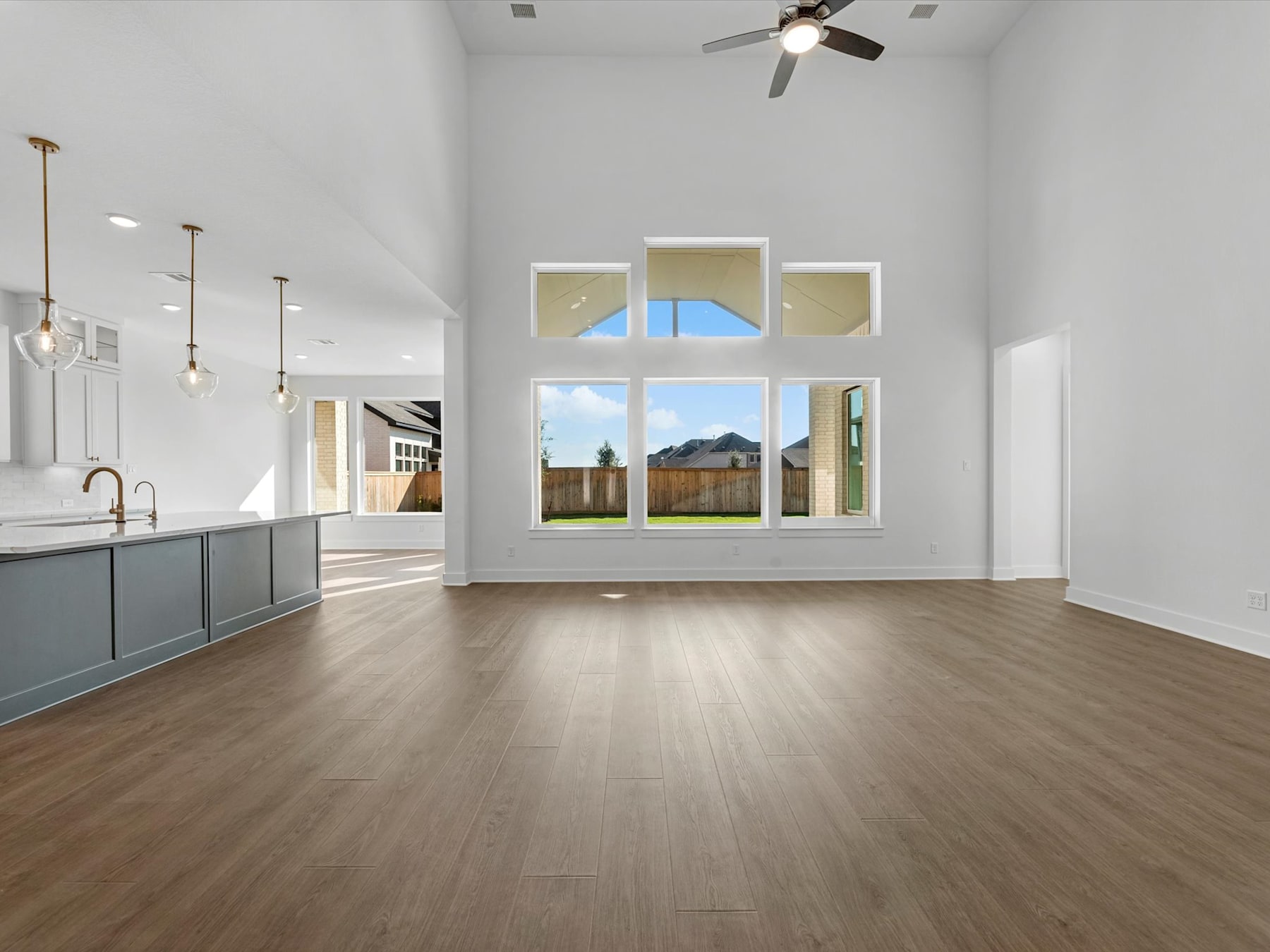 A spacious and bright open-plan living area with a kitchen visible in the background, featuring hardwood floors, large windows, and a ceiling fan.