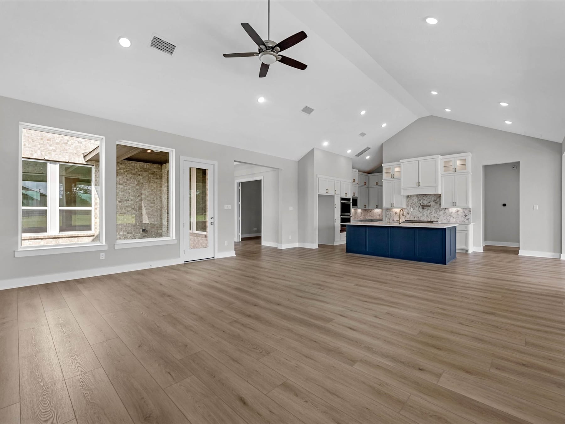 A spacious and modern open-plan living area with a vaulted ceiling, hardwood floors, and a kitchen with blue cabinetry visible in the background.
