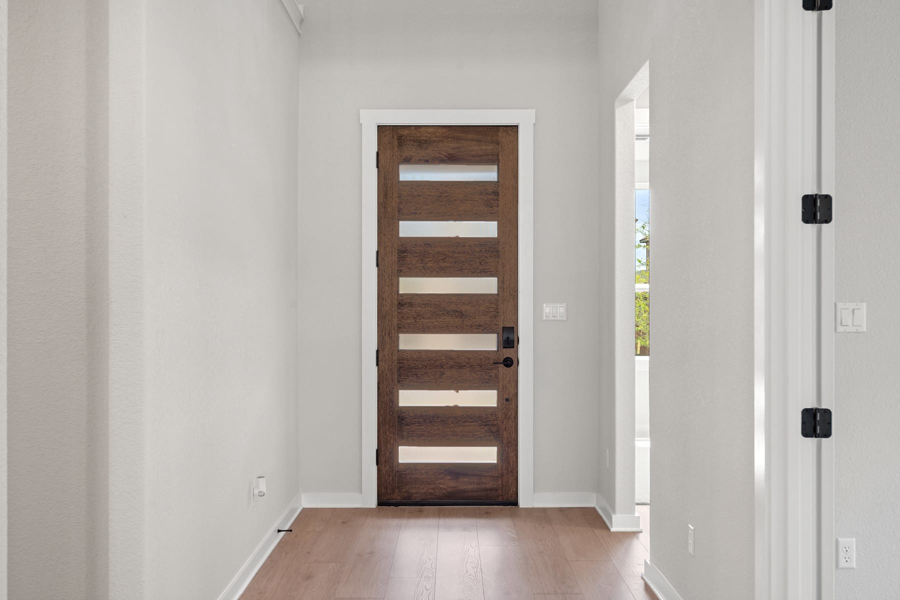 A wooden door with horizontal panels stands in a bright, white-walled hallway, leading to an open space beyond.