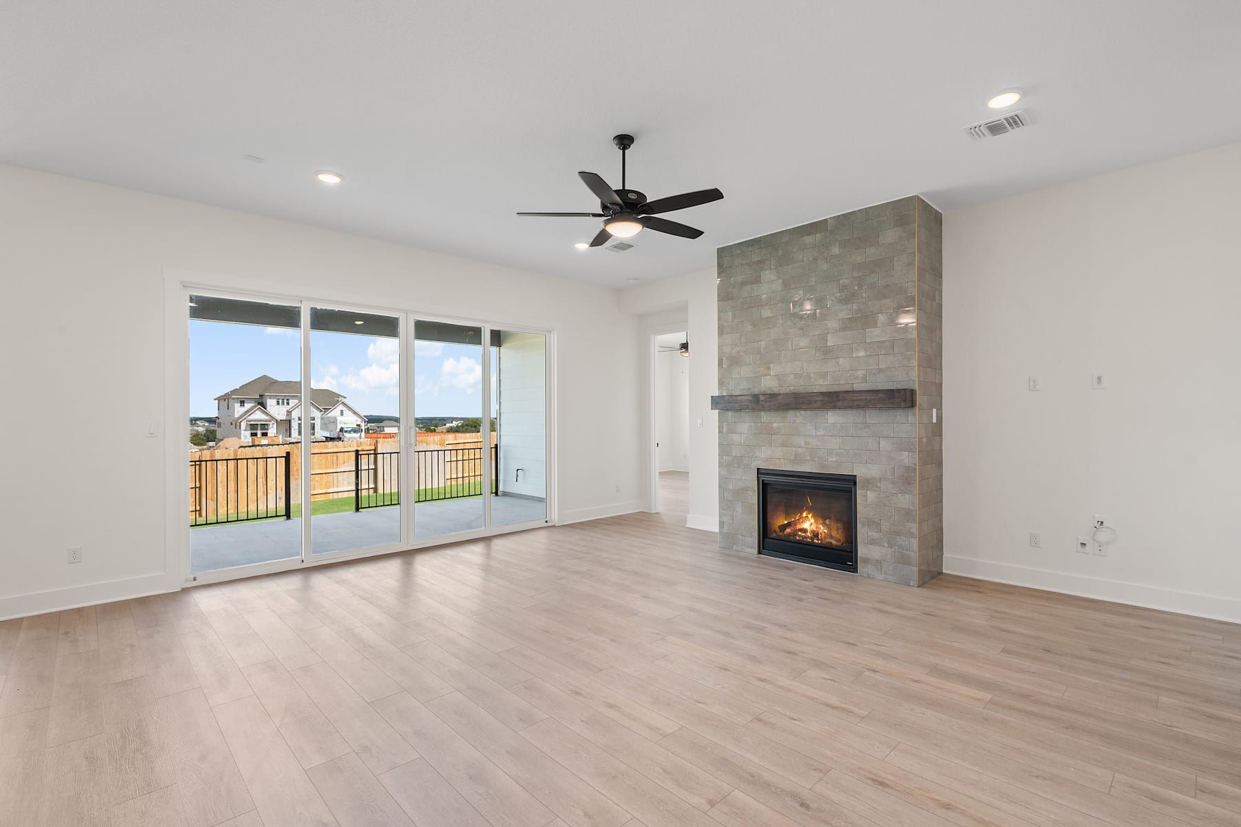 A spacious and bright living room with a fireplace, hardwood floors, and a ceiling fan, overlooking a backyard through large sliding glass doors.