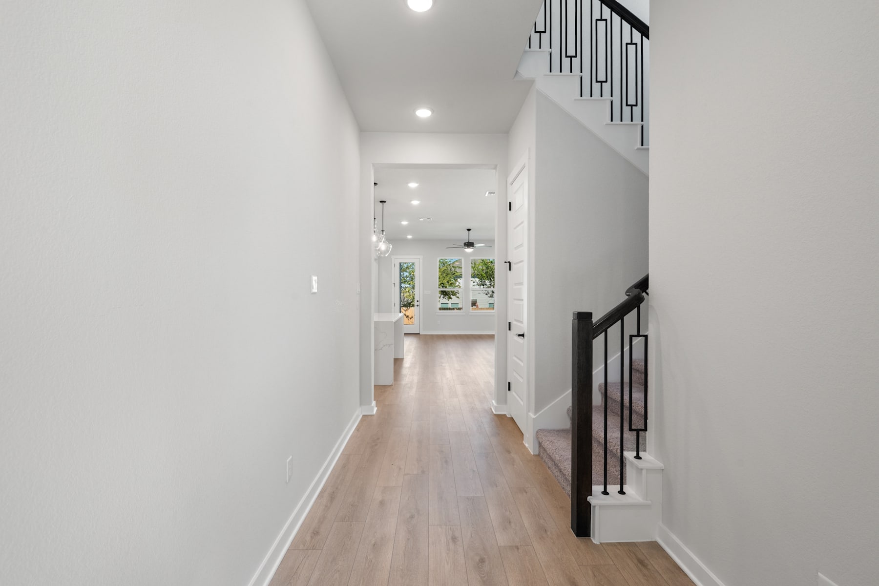 A bright, minimalist hallway with white walls, a wooden floor, and a window at the end, leading to a view of the outdoors.