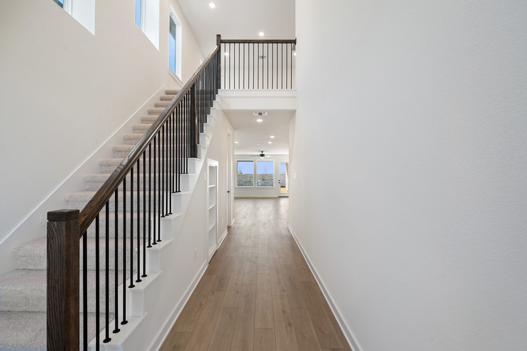 A bright, open hallway with a wooden staircase and railing leading up to a second floor, with a window at the end providing natural light.