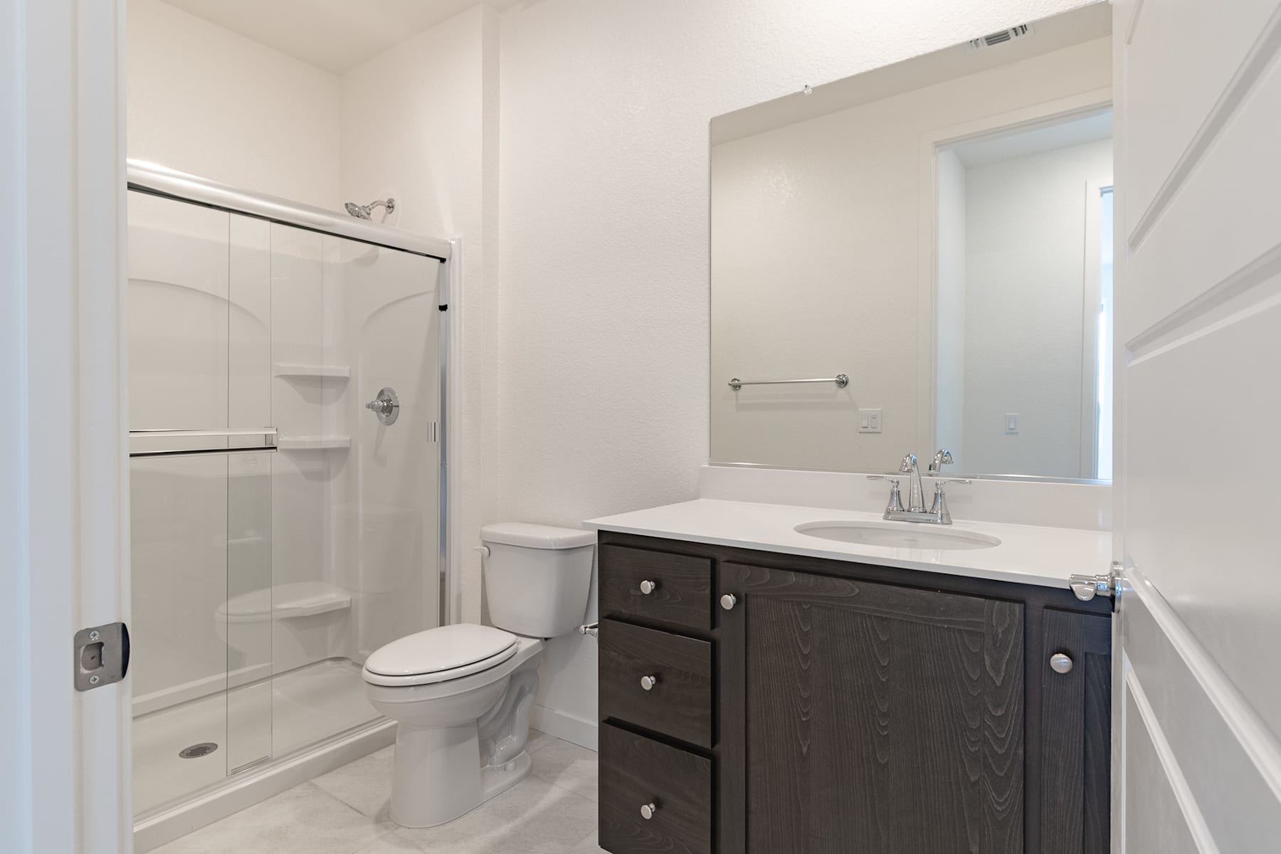 A modern bathroom with a dark vanity cabinet, a white sink, and a large mirror on the wall. The room has a clean, minimalist design with a shower enclosure visible in the background.