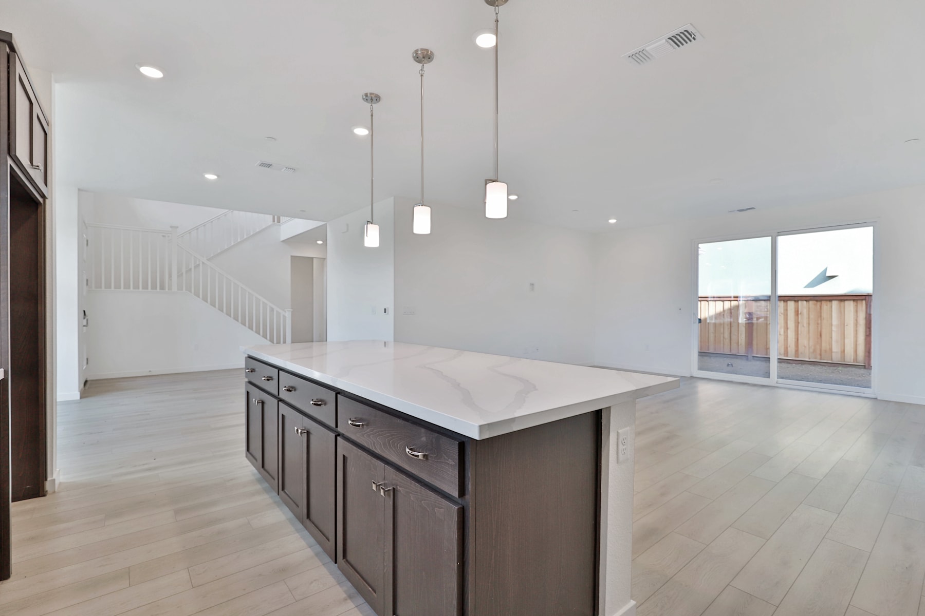A modern, open-concept kitchen with a large island countertop, dark cabinets, and pendant lighting fixtures, set against a bright, airy background with hardwood floors and a sliding glass door leading to an outdoor area.