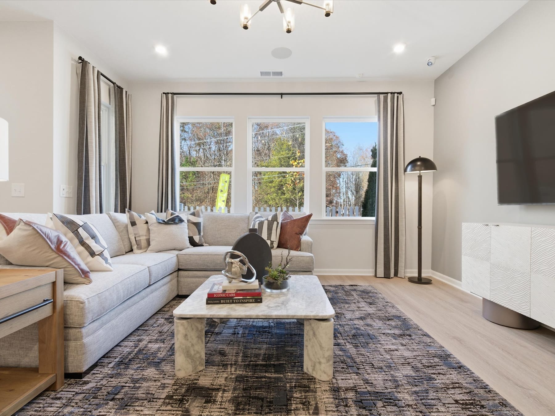 A cozy and inviting living room with a large sectional sofa, a wooden coffee table, and a chandelier overhead, surrounded by large windows that provide a view of the outdoor scenery.