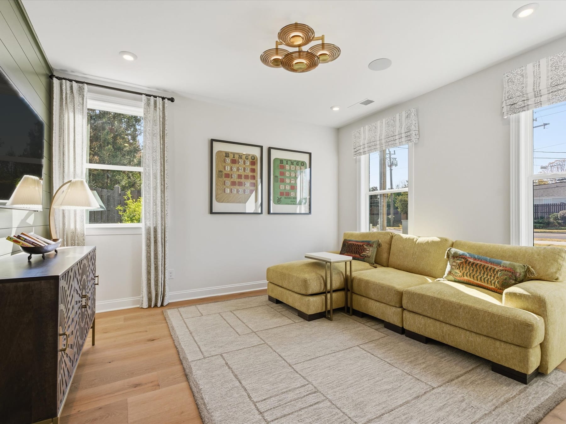 A cozy living room with a yellow sectional sofa, a patterned area rug, and a modern chandelier overhead, surrounded by framed artwork on the walls.