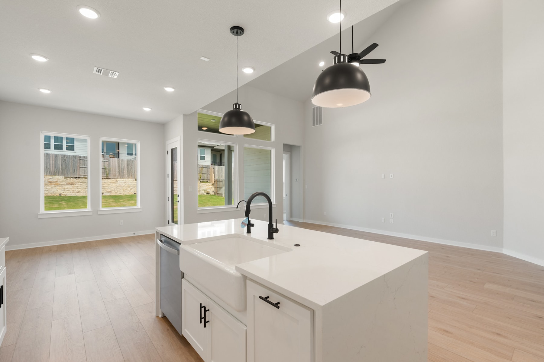 A modern kitchen with white cabinets, a farmhouse-style sink, and pendant lights hanging above the counter. The room has large windows that provide natural light and a view of the outdoor space.