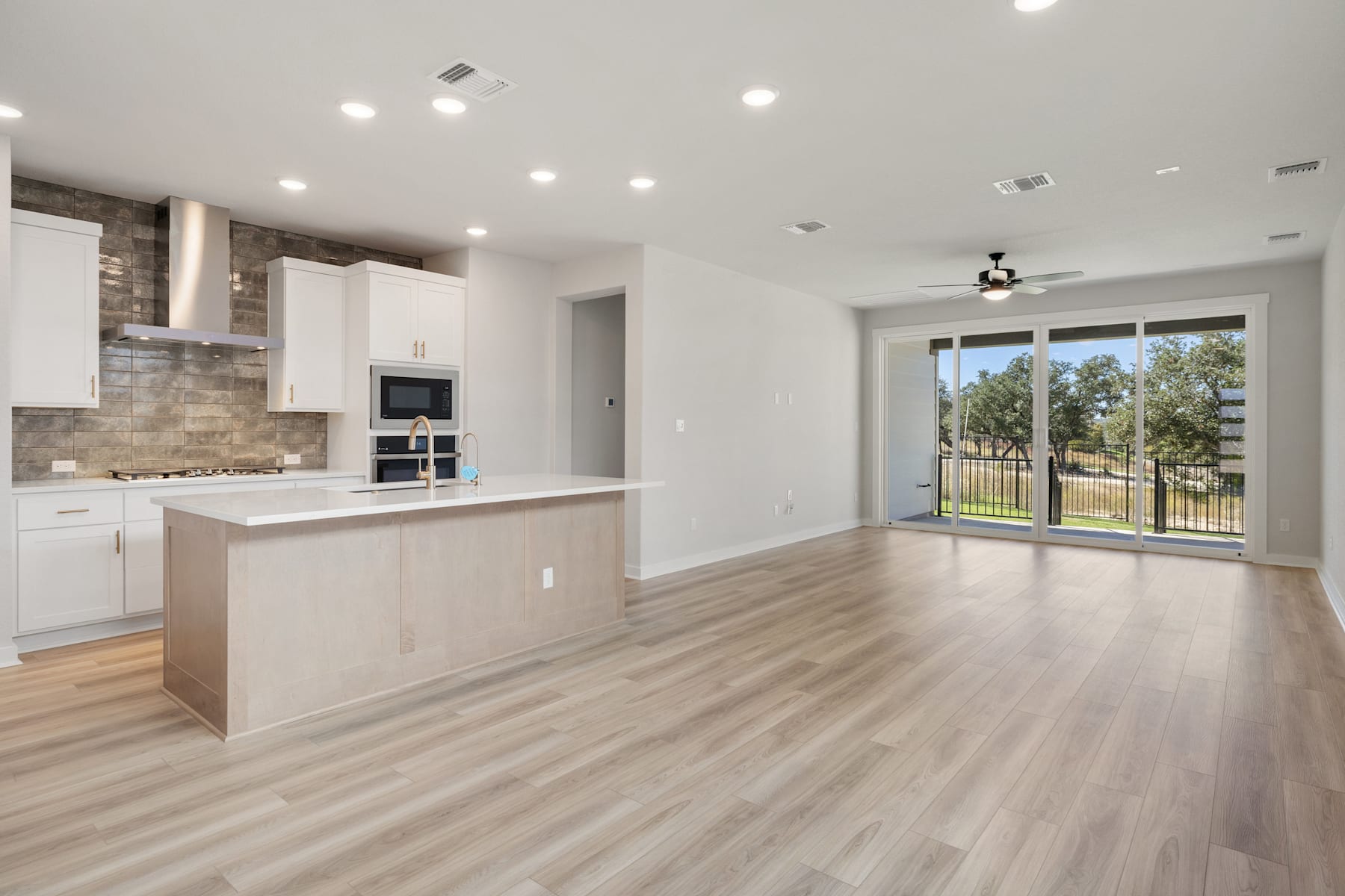 A modern, open-concept kitchen and living room with white cabinets, a tile backsplash, and hardwood floors, overlooking a scenic outdoor view through a large window.