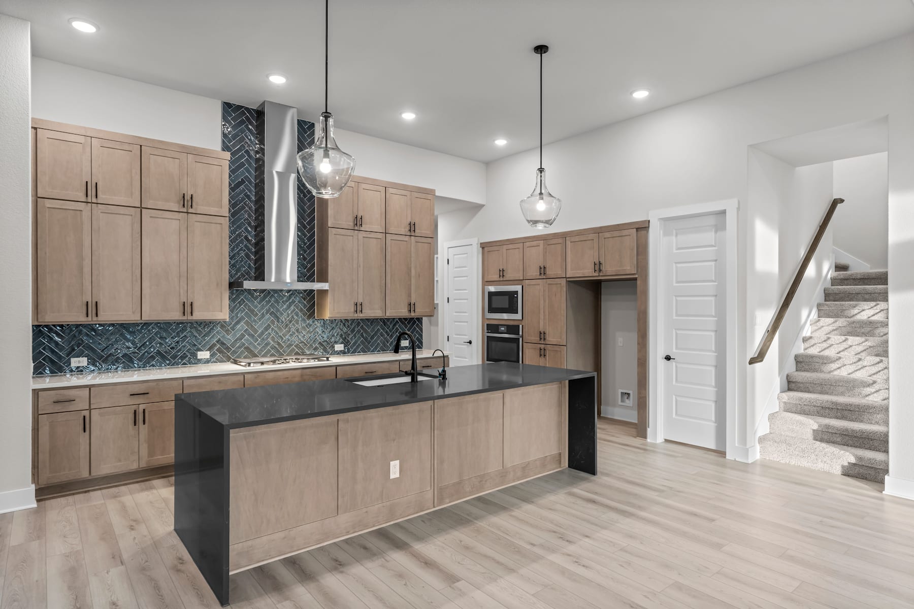 A modern, open-concept kitchen with light wood cabinets, a black countertop, and pendant lighting fixtures, set against a backdrop of white walls and hardwood floors.