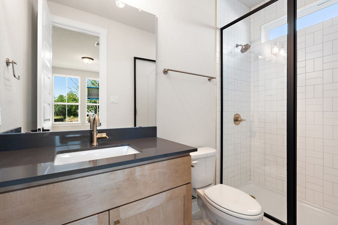 A modern bathroom with a wooden vanity, a black countertop, and a glass-enclosed shower in the background, overlooking a window with a view of the outdoors.