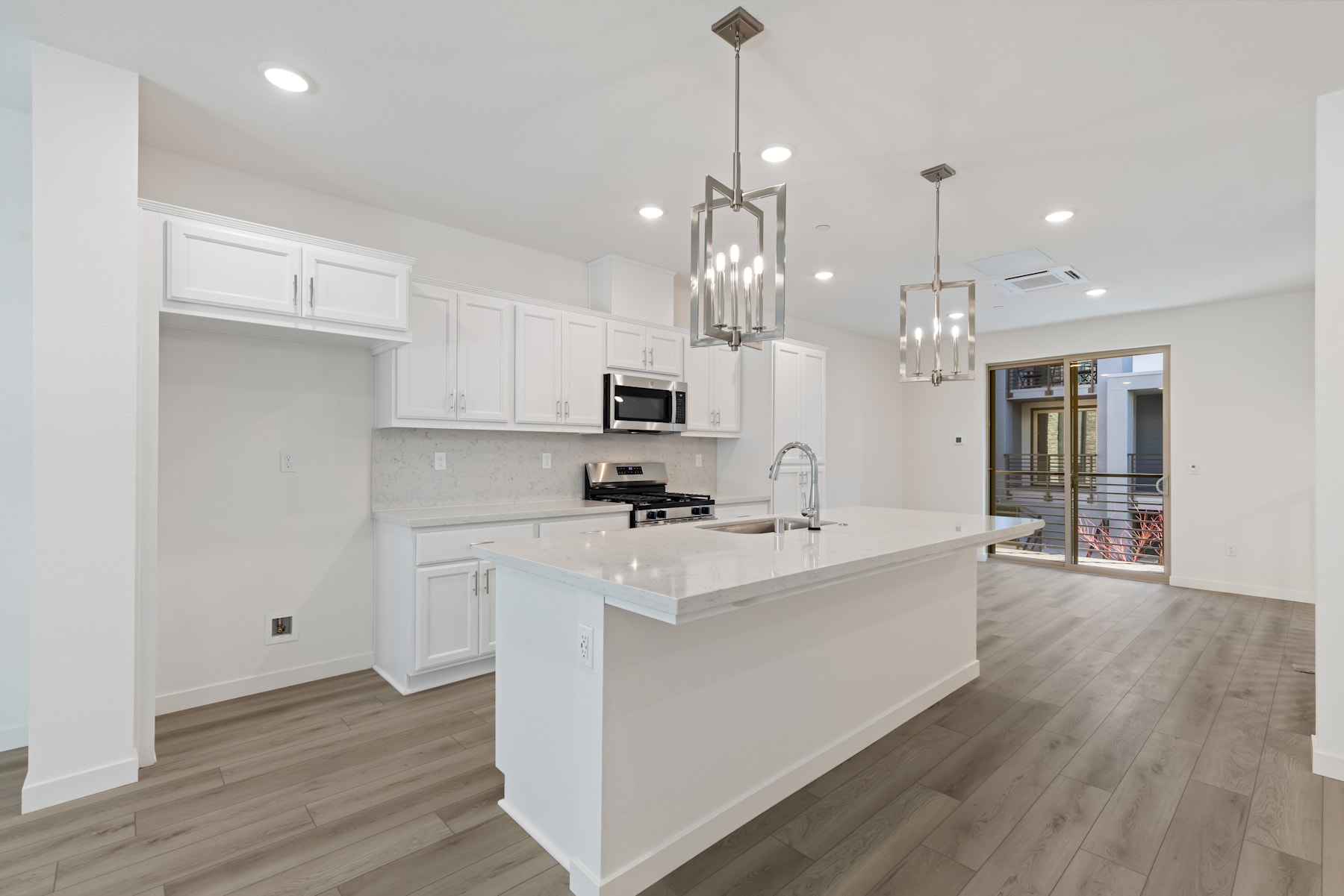 A modern, open-concept kitchen with white cabinets, a large island, and sleek pendant lighting fixtures, set against a backdrop of hardwood floors and a hallway leading to another room.