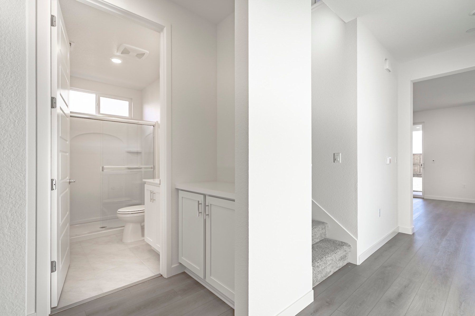A bright, minimalist hallway with a partially visible bathroom doorway, leading to a well-lit and spacious interior.