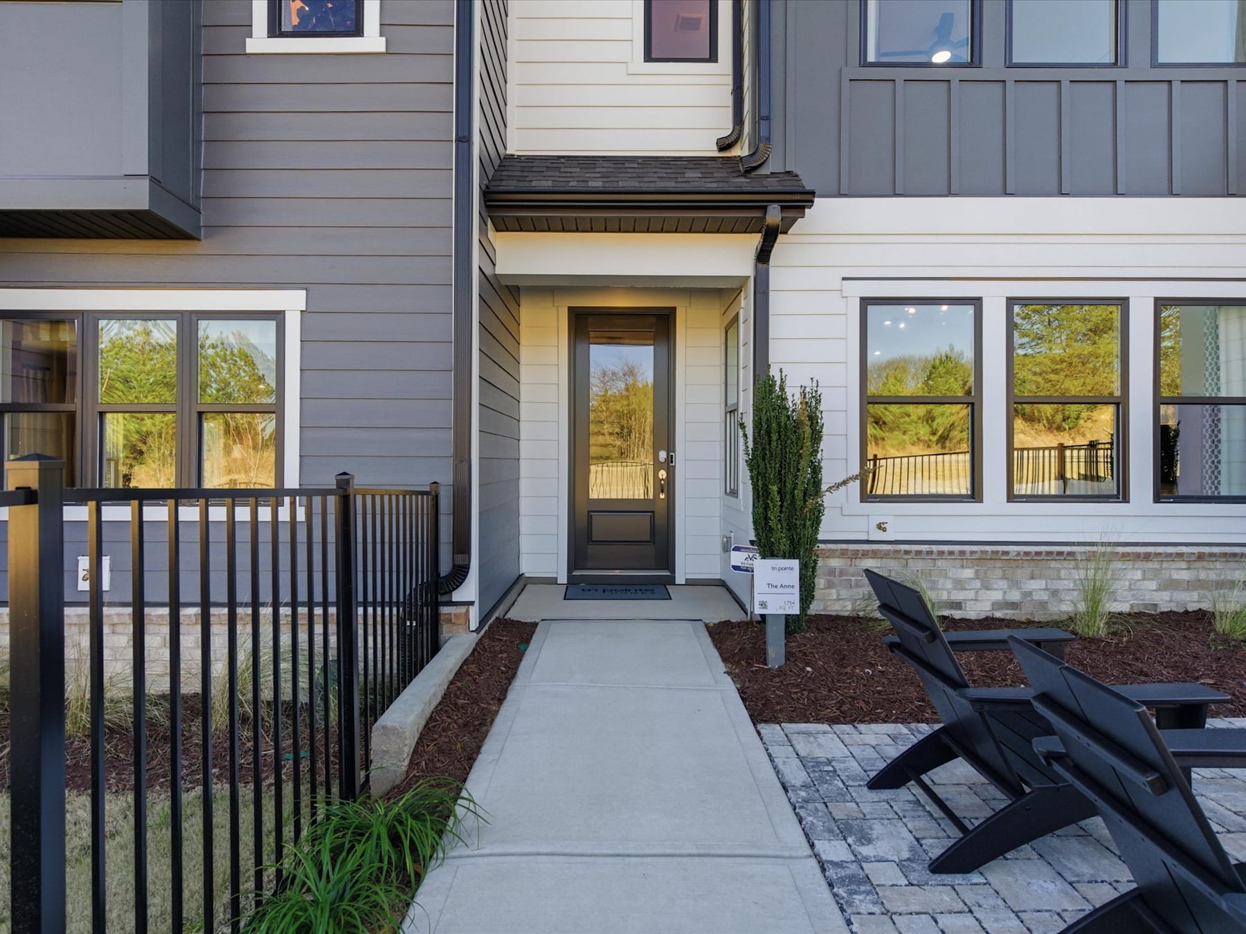A modern, two-story residential building with a paved walkway leading to the front entrance, surrounded by landscaping and a metal railing.