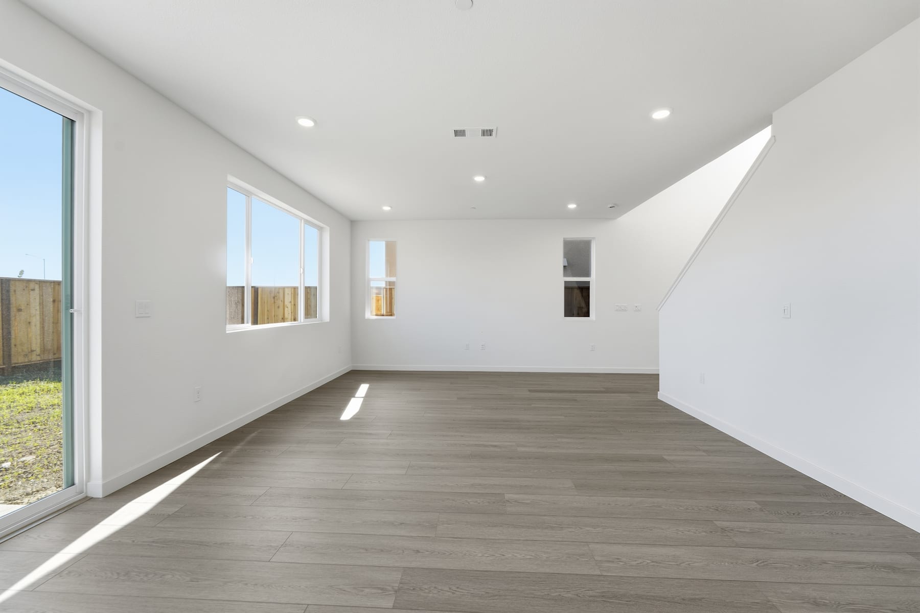 A spacious, well-lit room with a wooden floor and large windows overlooking a backyard.