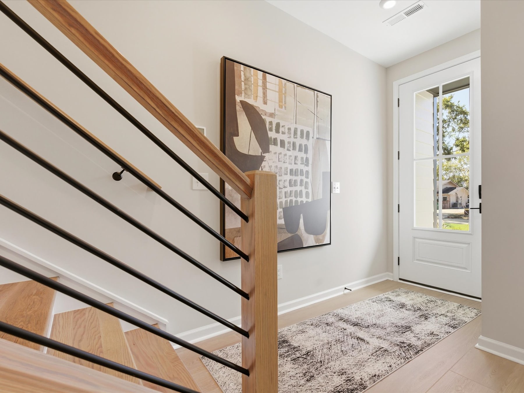 A modern and minimalist entryway with a wooden staircase, a framed abstract artwork on the wall, and a patterned rug on the floor, leading to a bright and airy space beyond.