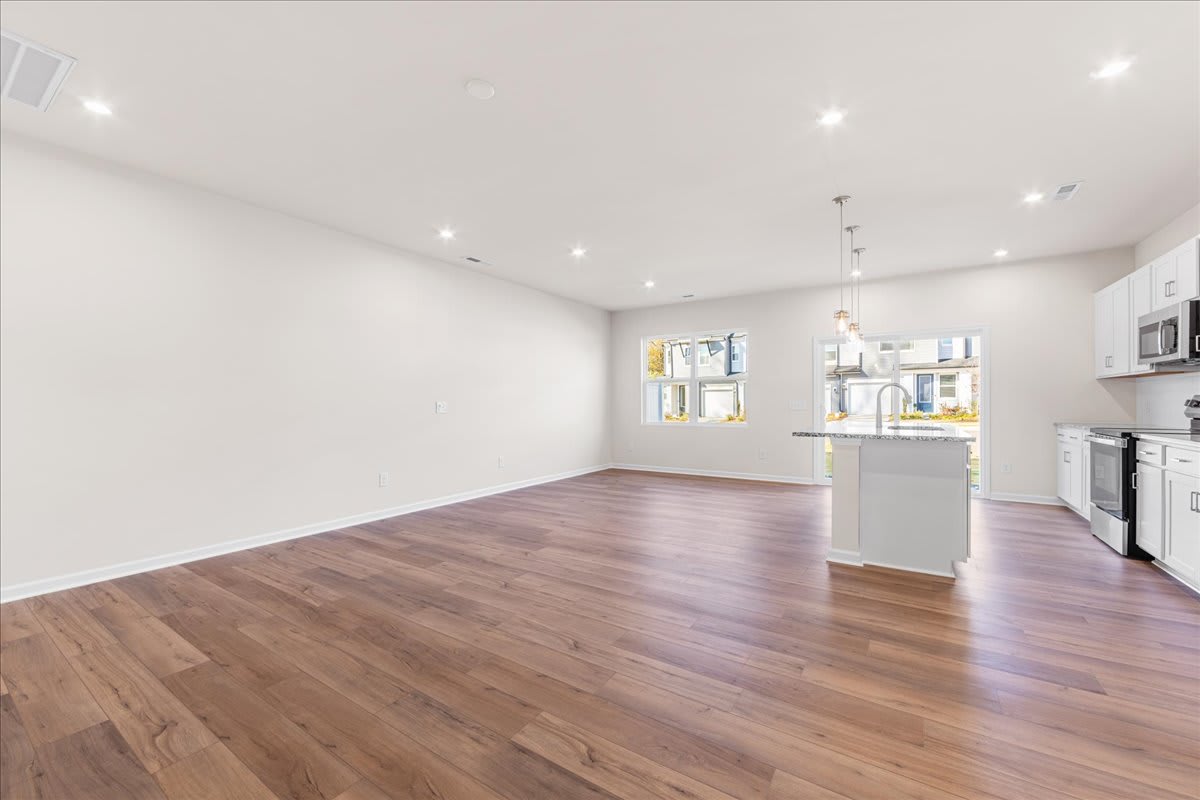A spacious, well-lit room with hardwood floors, white walls, and recessed lighting fixtures. The room appears to be an open-concept living or dining area, with a kitchen visible in the background.