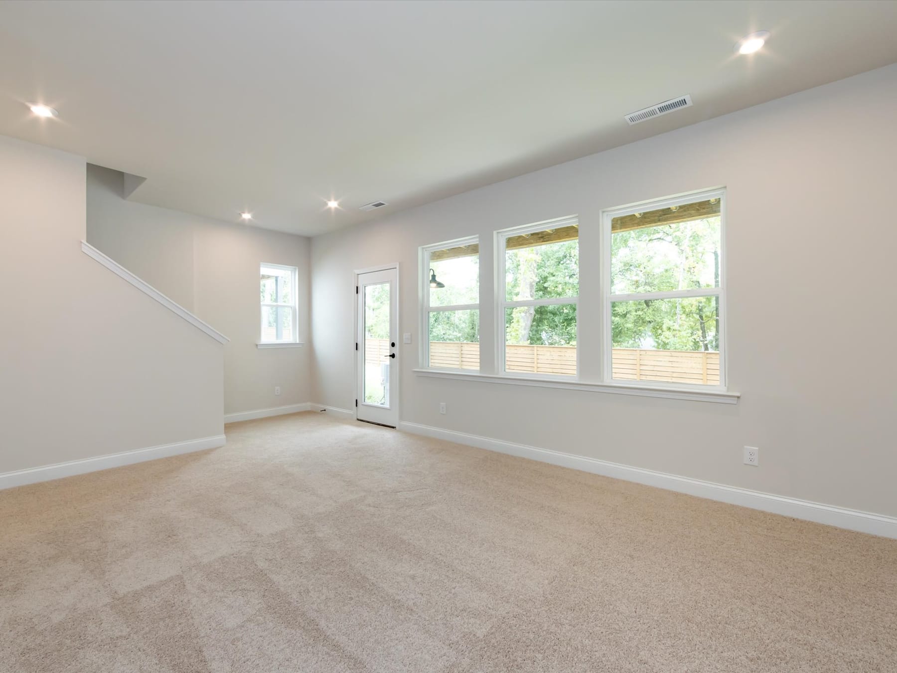A spacious, well-lit room with large windows overlooking a lush, green outdoor area, featuring a neutral-toned carpet and clean, minimalist decor.
