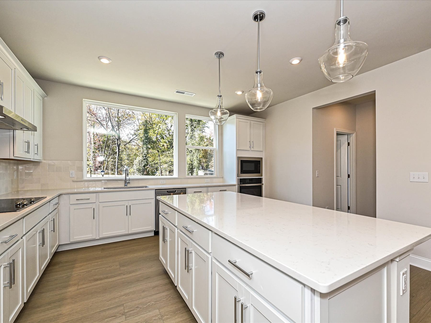A modern, bright kitchen with white cabinets, a large island, and pendant lighting, set against a backdrop of trees visible through the windows.