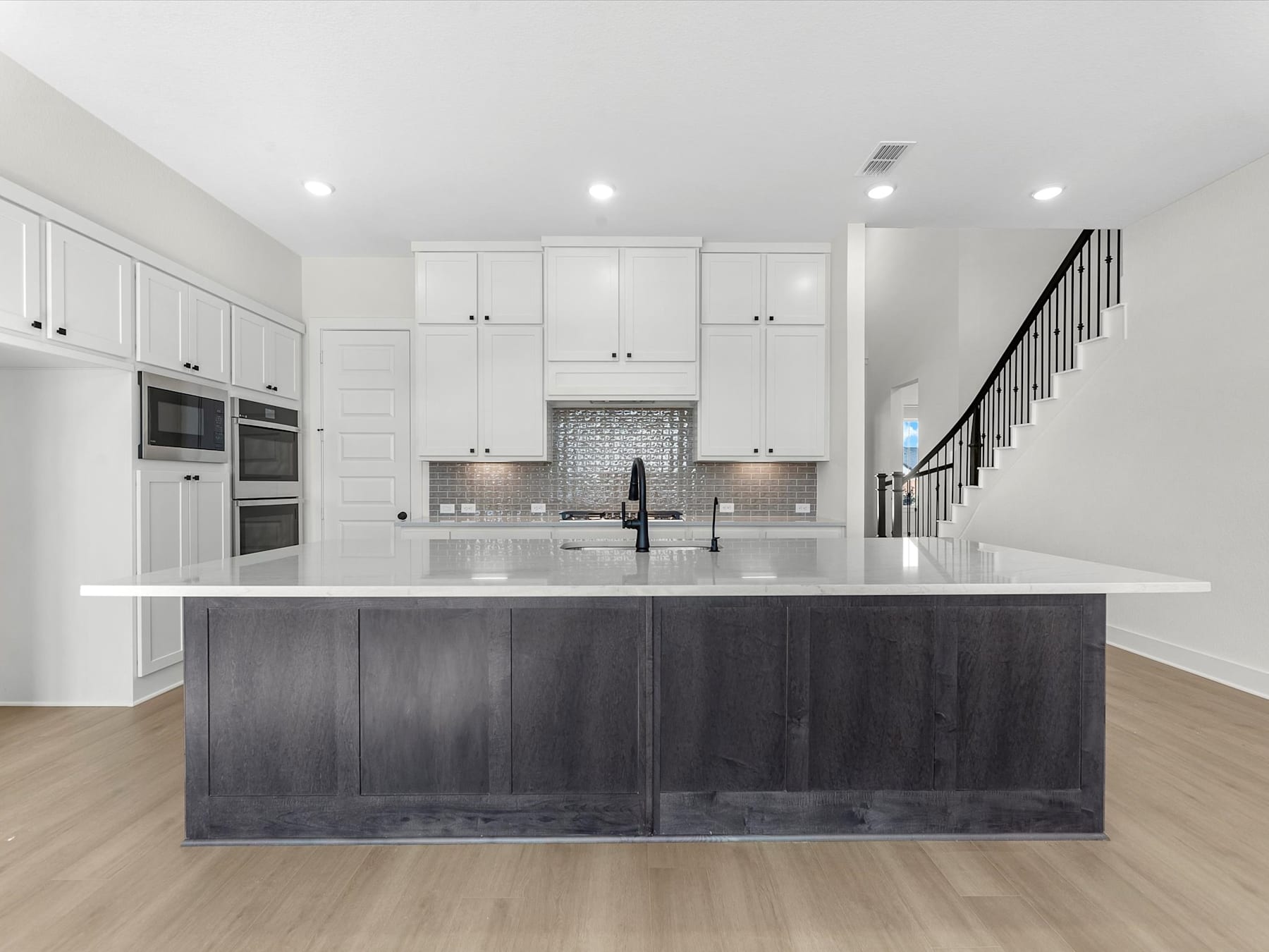 A modern, open-concept kitchen with white cabinets, a large dark-colored island, and a brick backsplash, set against a bright and airy background.