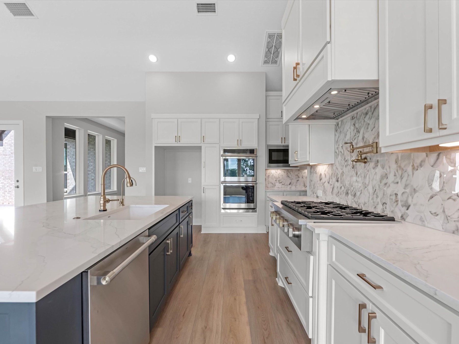 A modern, open-concept kitchen with white cabinets, marble backsplash, and stainless steel appliances, set against a hardwood floor.